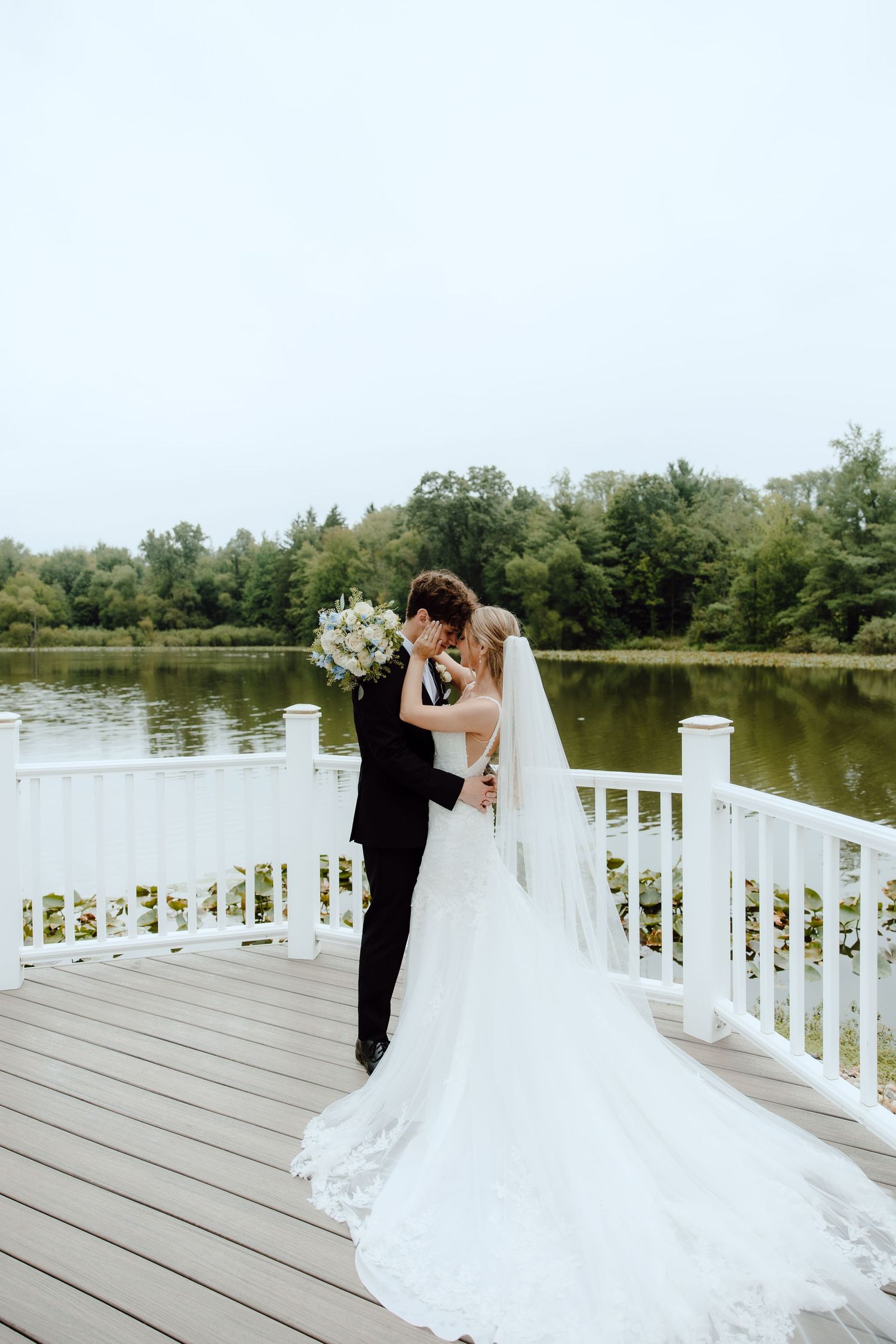 Bride and groom share romantic kiss on white waterfront deck surrounded by lush green trees.