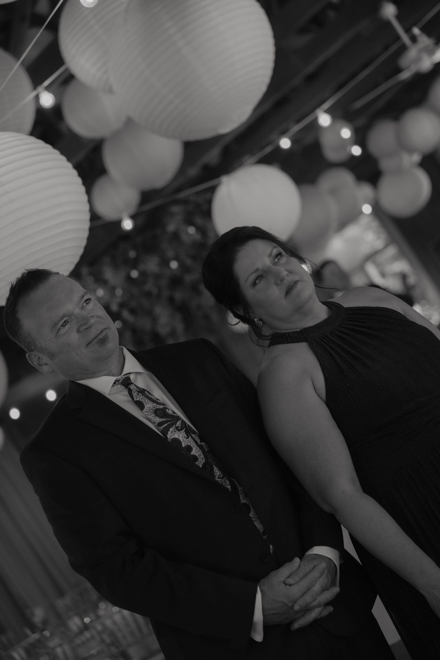 Black and white photo of an elegant couple holding hands under decorative paper lanterns at a wedding reception.