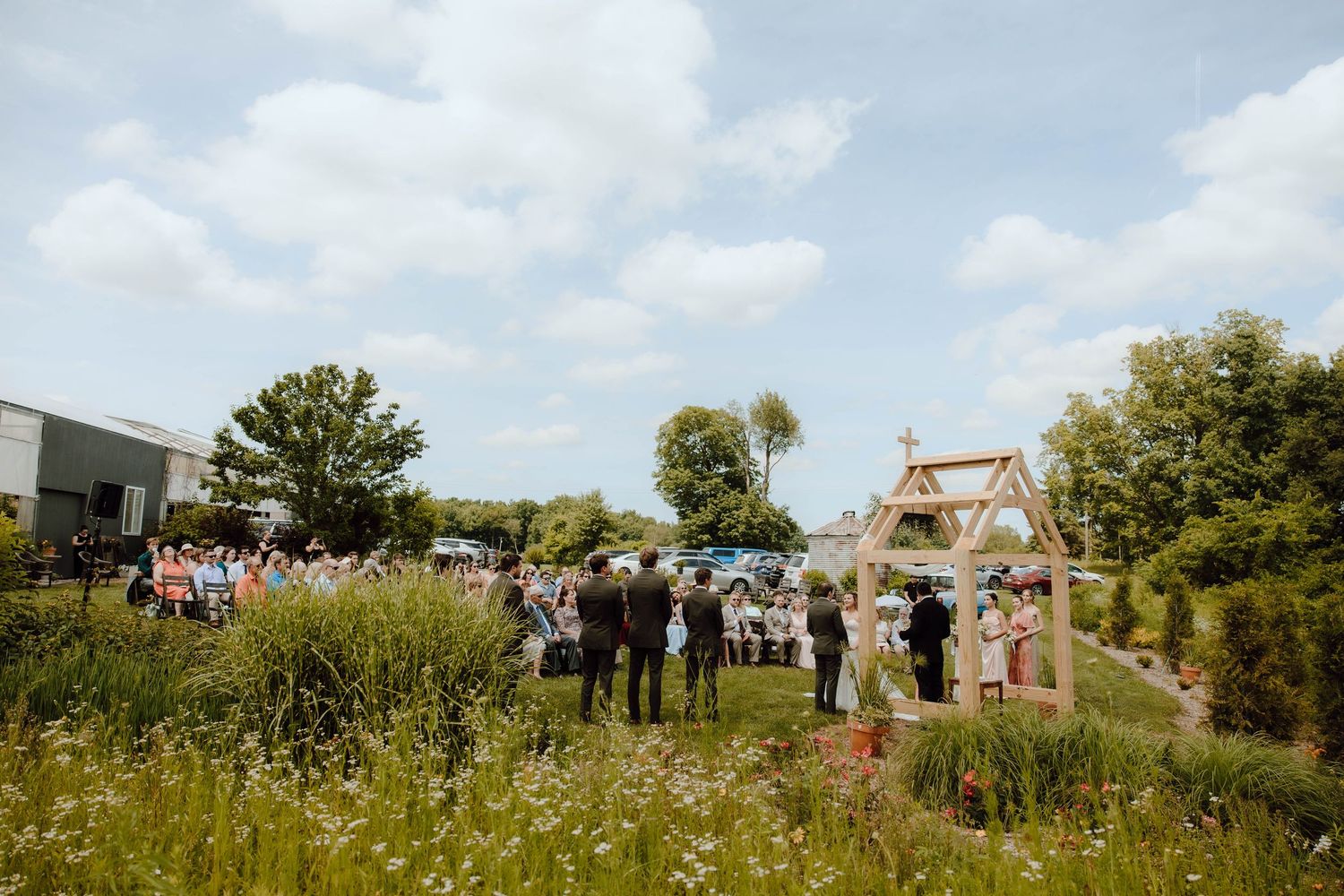 Outdoor wedding ceremony in a garden setting with guests gathered around a wooden arch under a partly cloudy sky.