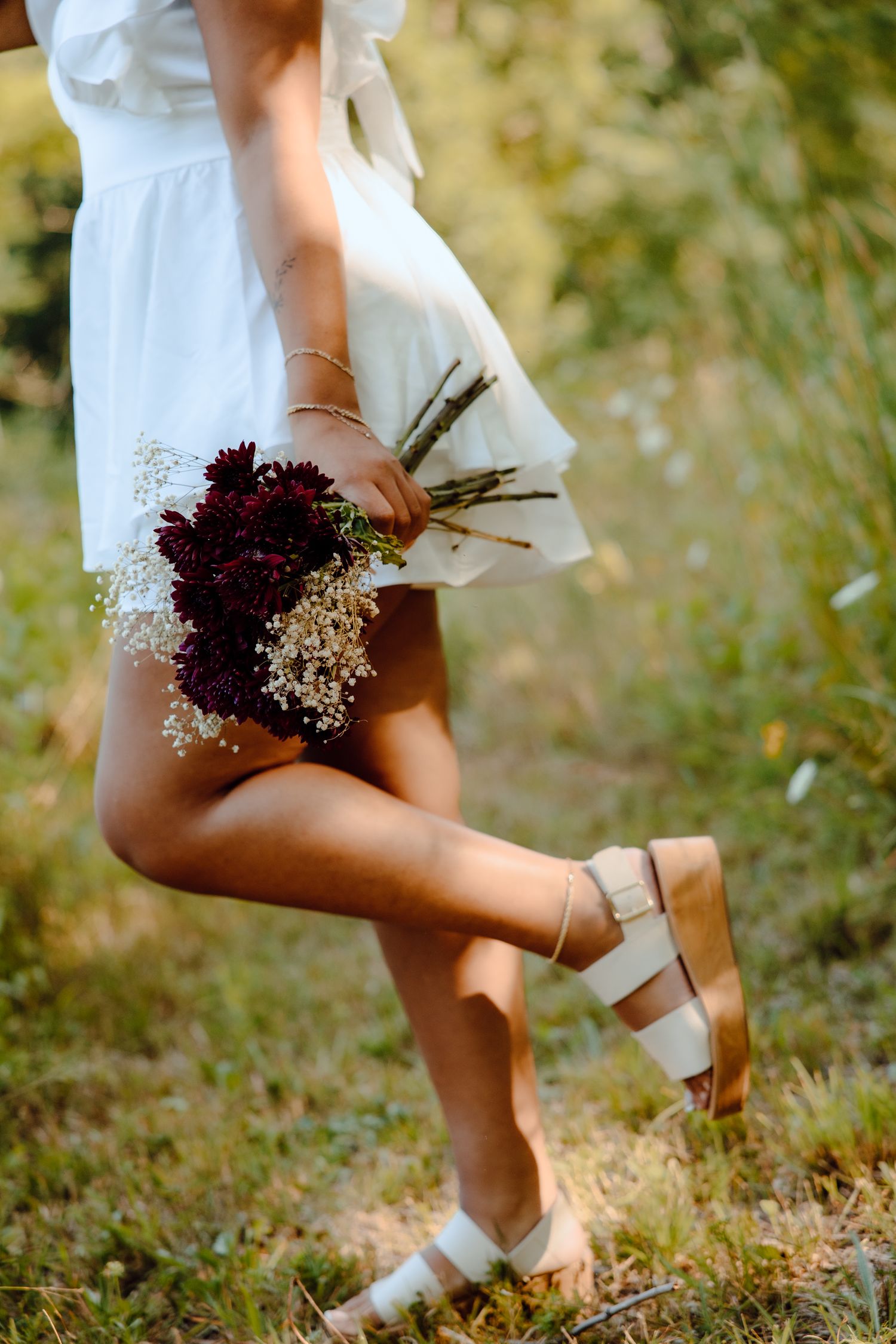 Close-up of white dress and burgundy bouquet against grassy summer field.