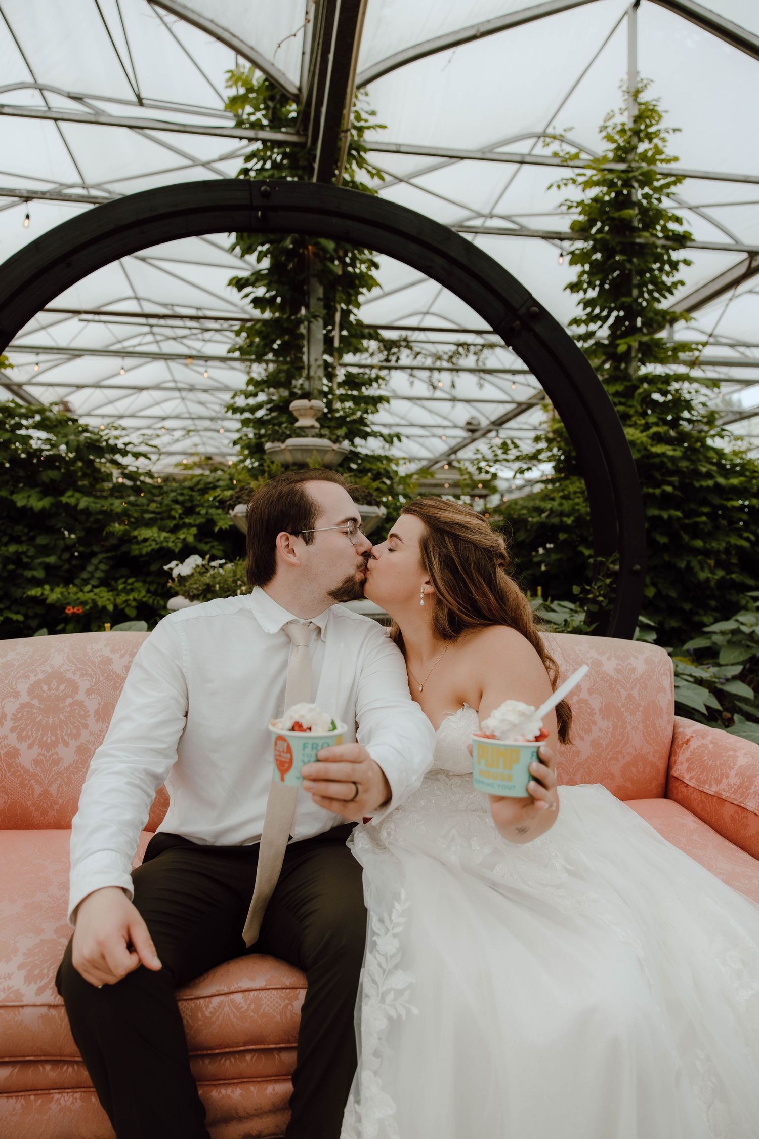 Newlyweds kissing on a pink sofa in a greenhouse, holding ice cream cups in wedding attire.