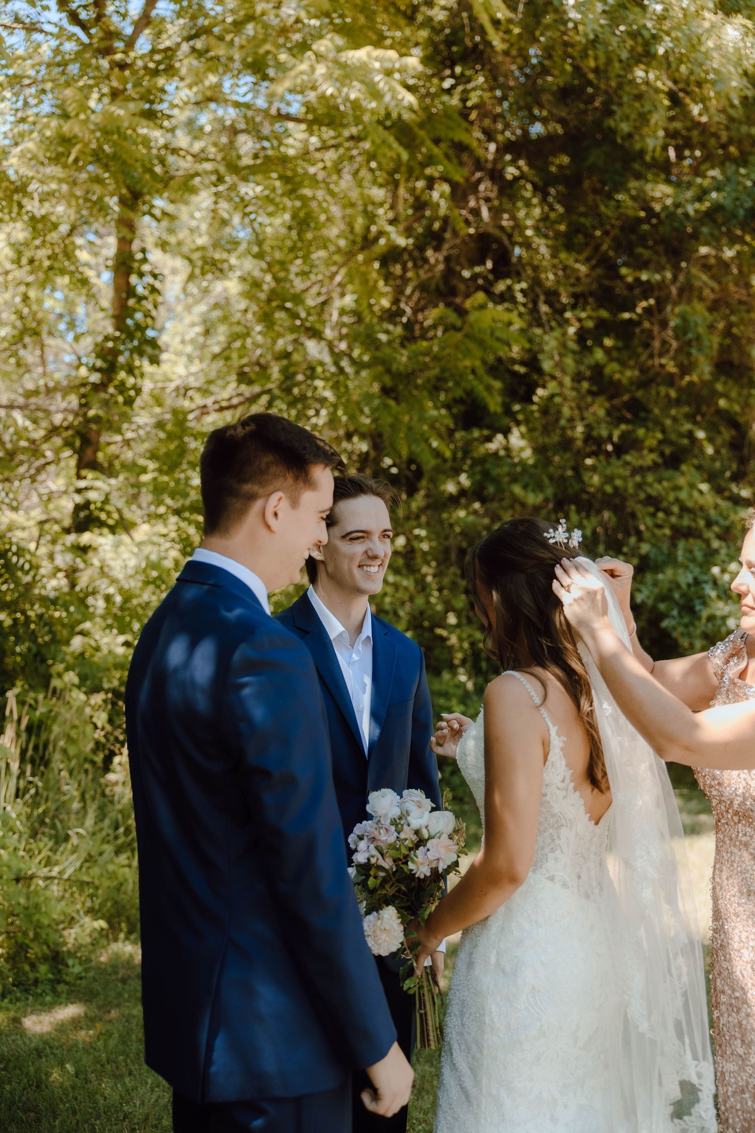 Outdoor wedding ceremony with groom in navy suit, smiling man, and bride in lace dress holding white floral bouquet.