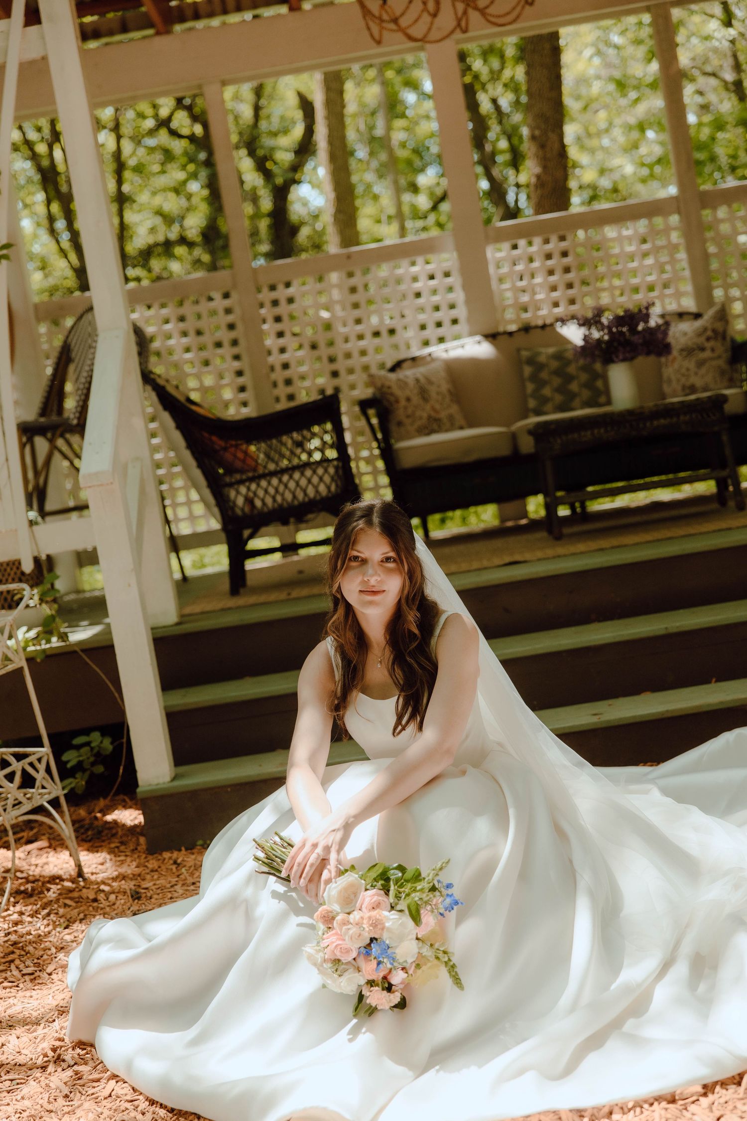 Bride in white gown sits gracefully outdoors holding pastel bouquet near rustic gazebo with lush green trees.