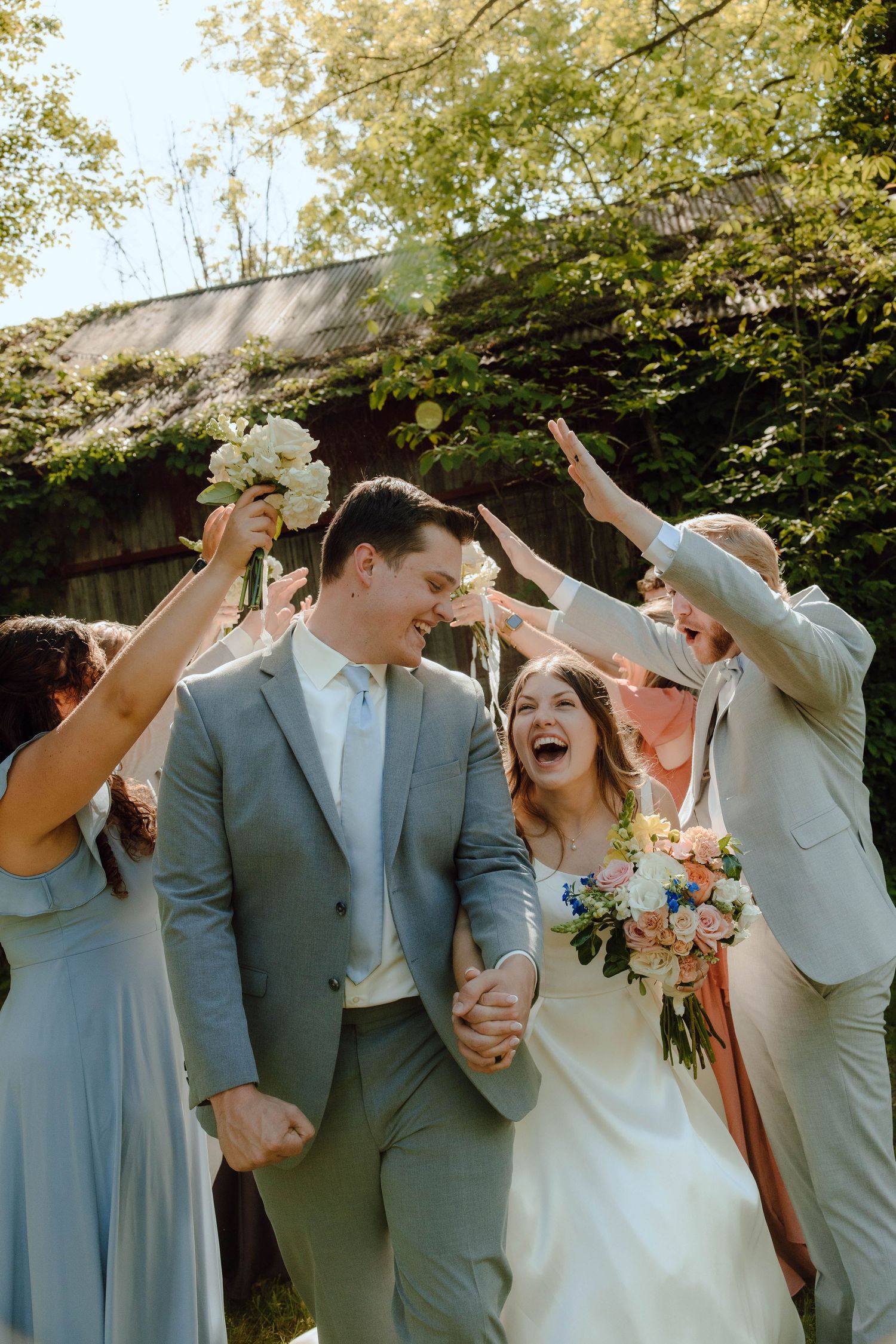 A joyful bride and groom walk hand-in-hand under raised bouquets as their wedding party celebrates outdoors.