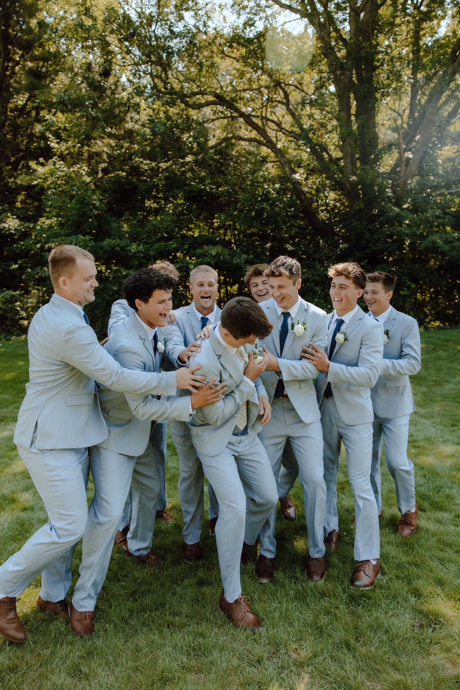 A groom in a light blue suit laughs with his groomsmen in matching suits on a green lawn surrounded by trees.