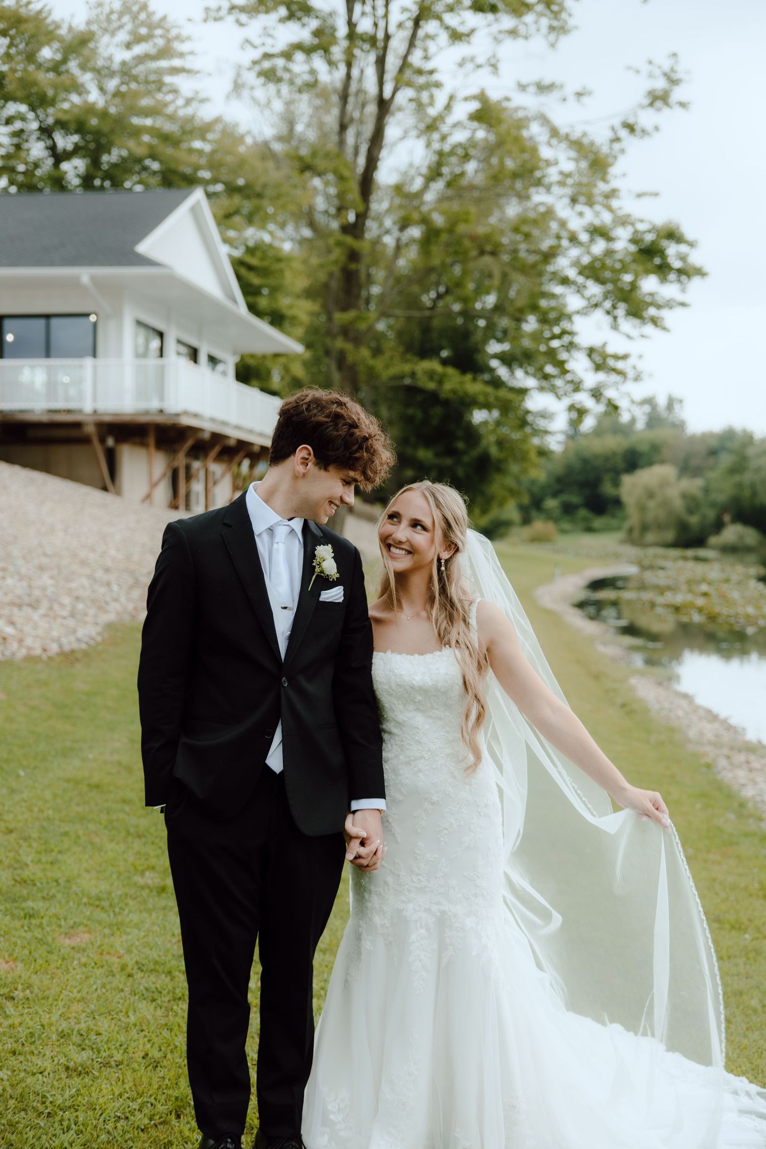 Smiling bride and groom walk hand-in-hand along a pond near a white venue building on their wedding day.