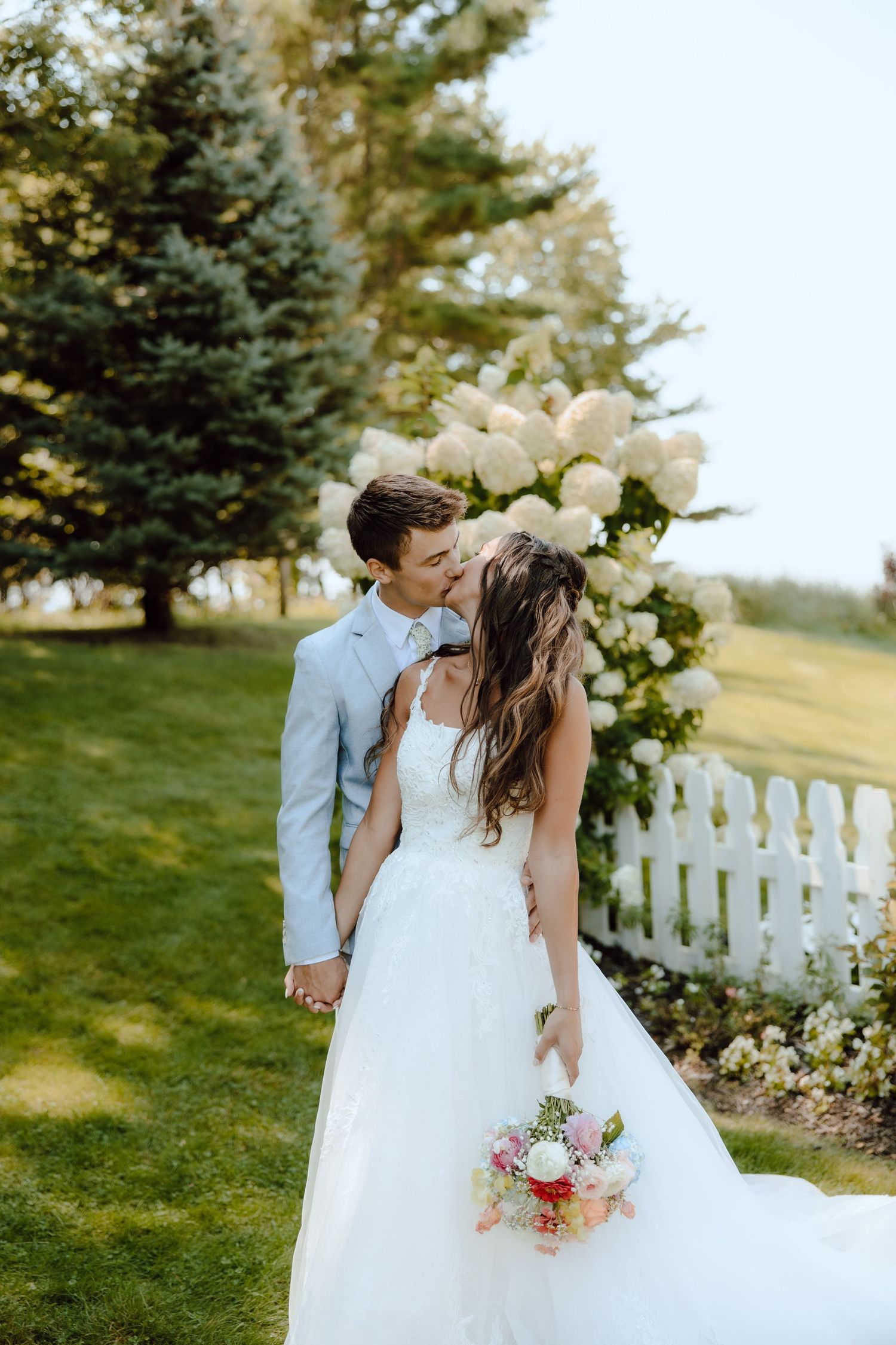 A groom in a light blue suit kisses his bride in a white gown holding a bouquet near a white picket fence.