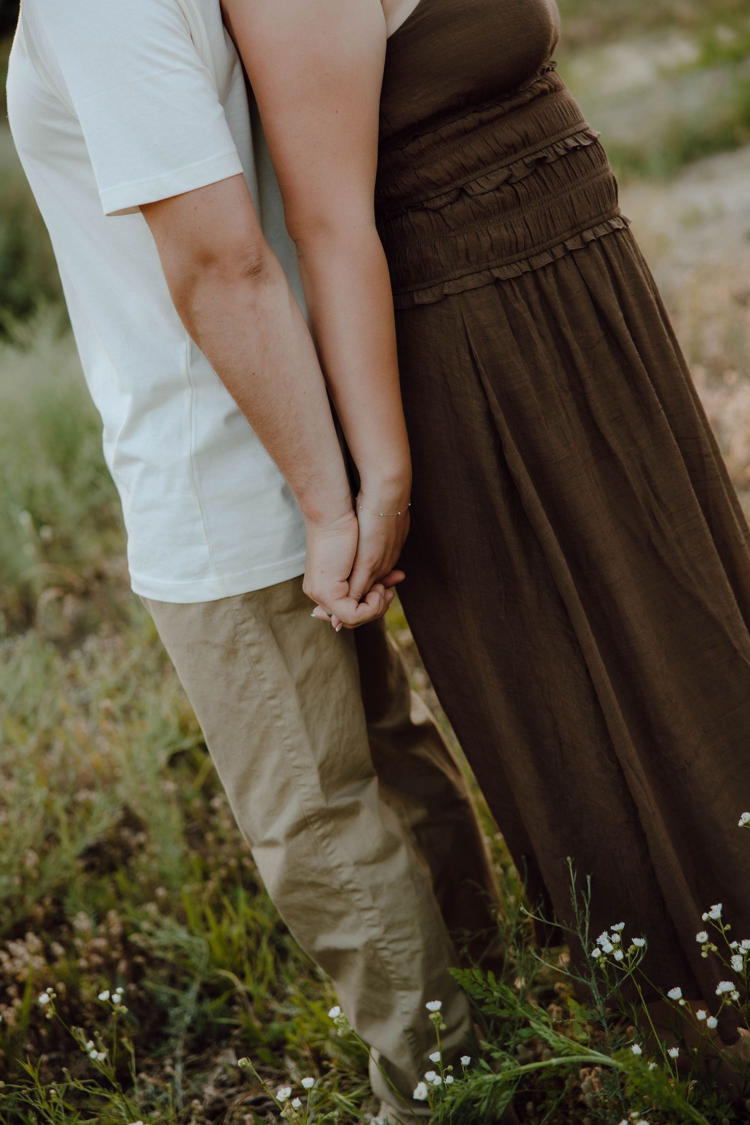 Couple holding hands outdoors, man in white shirt and khaki pants, woman in olive green maxi dress, standing in a field.