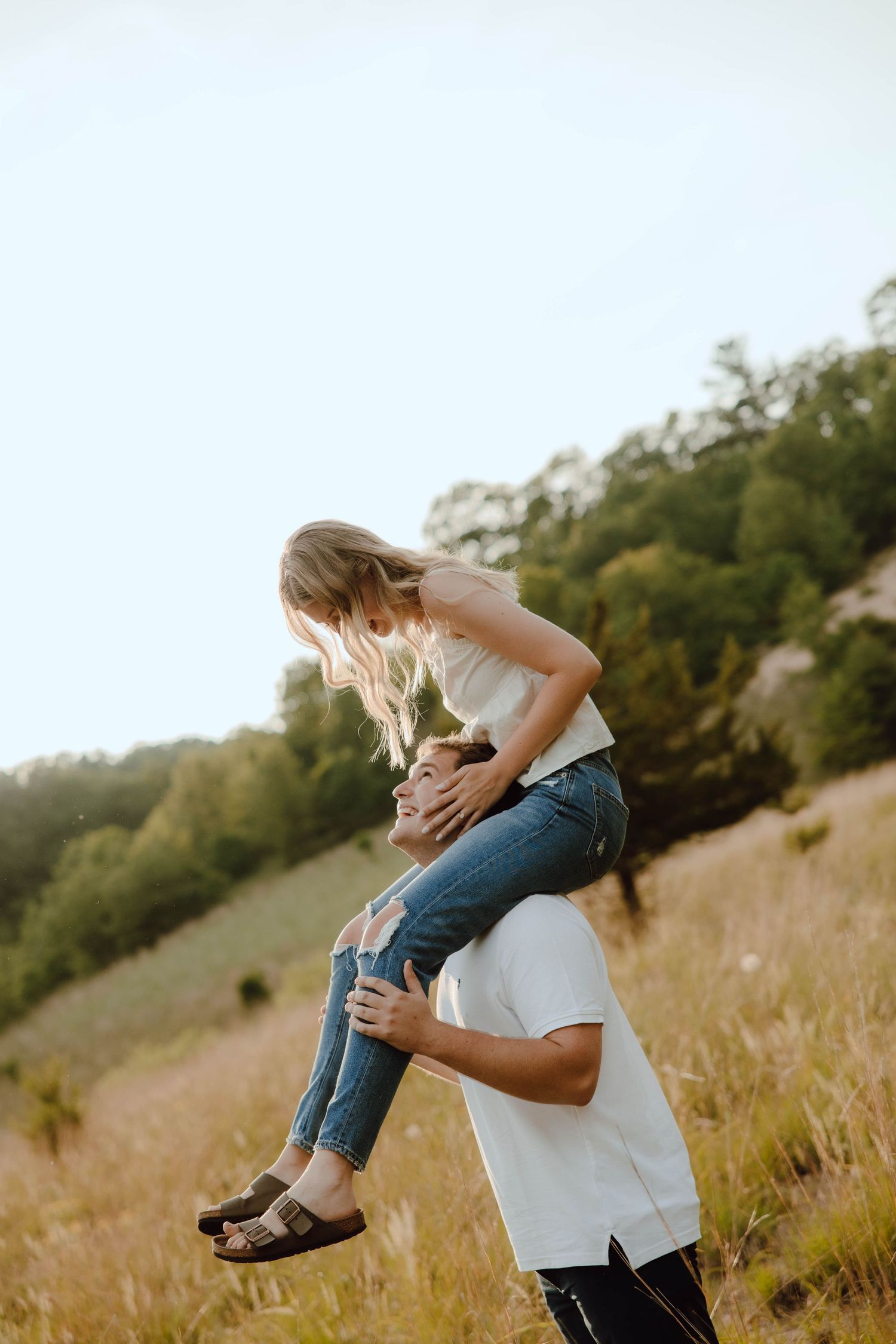 A man lifts a woman onto his shoulders in a sunny field, both smiling joyfully with green hills in the background.