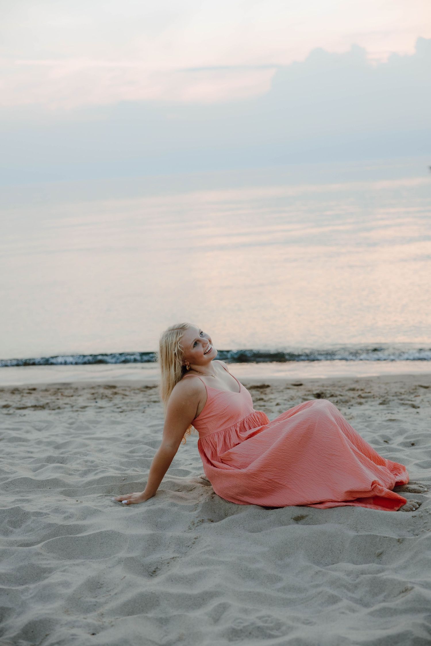 Blonde woman in pink maxi dress sitting on sandy beach at sunset with calm ocean in background.