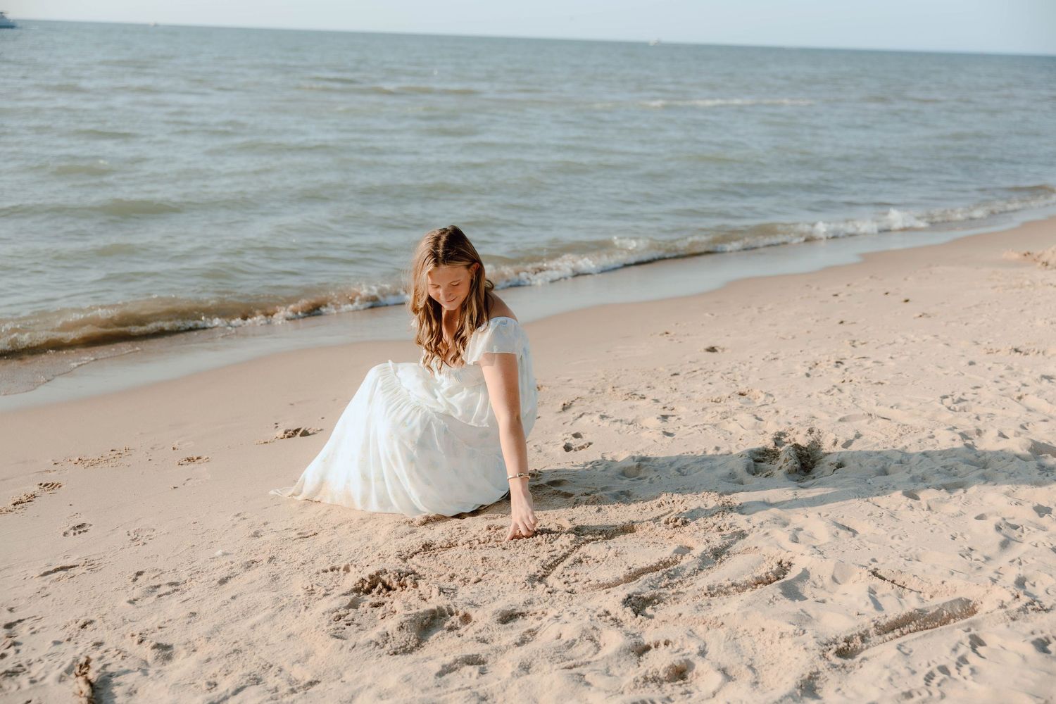 A young woman in a white dress sits on a sandy beach shore, playing in the sand near the ocean waves.