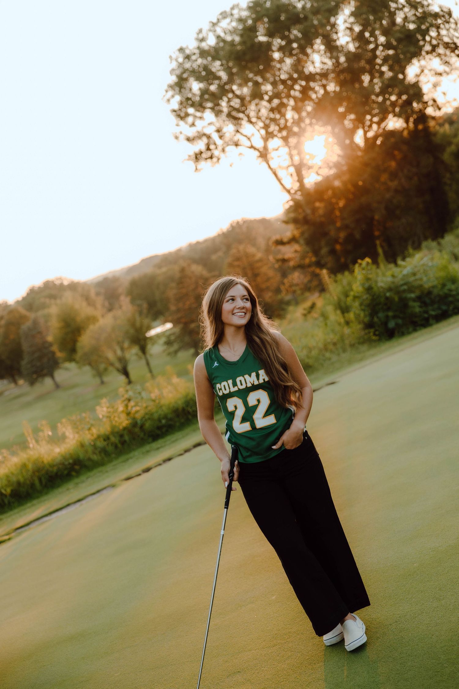 Young woman in green Coloma #22 jersey holds golf club on sunny golf course at golden hour.