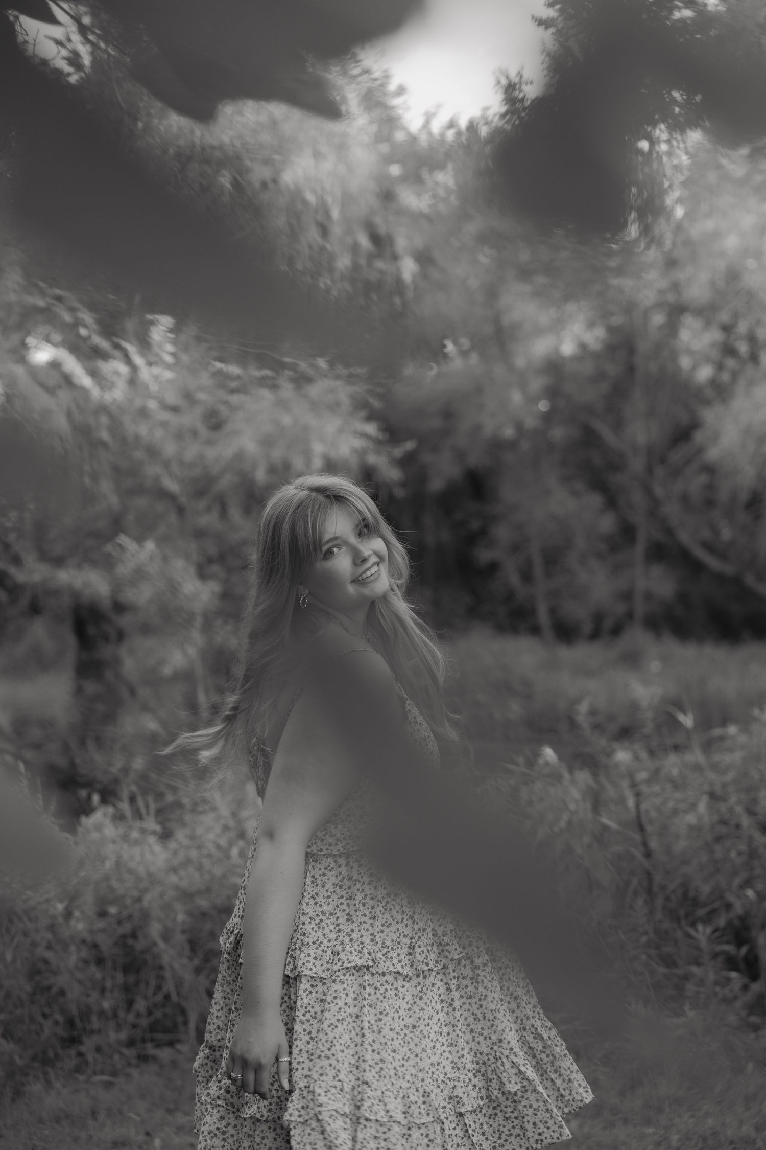 Black and white photo of a smiling young woman in a floral dress twirling outdoors among lush green trees.