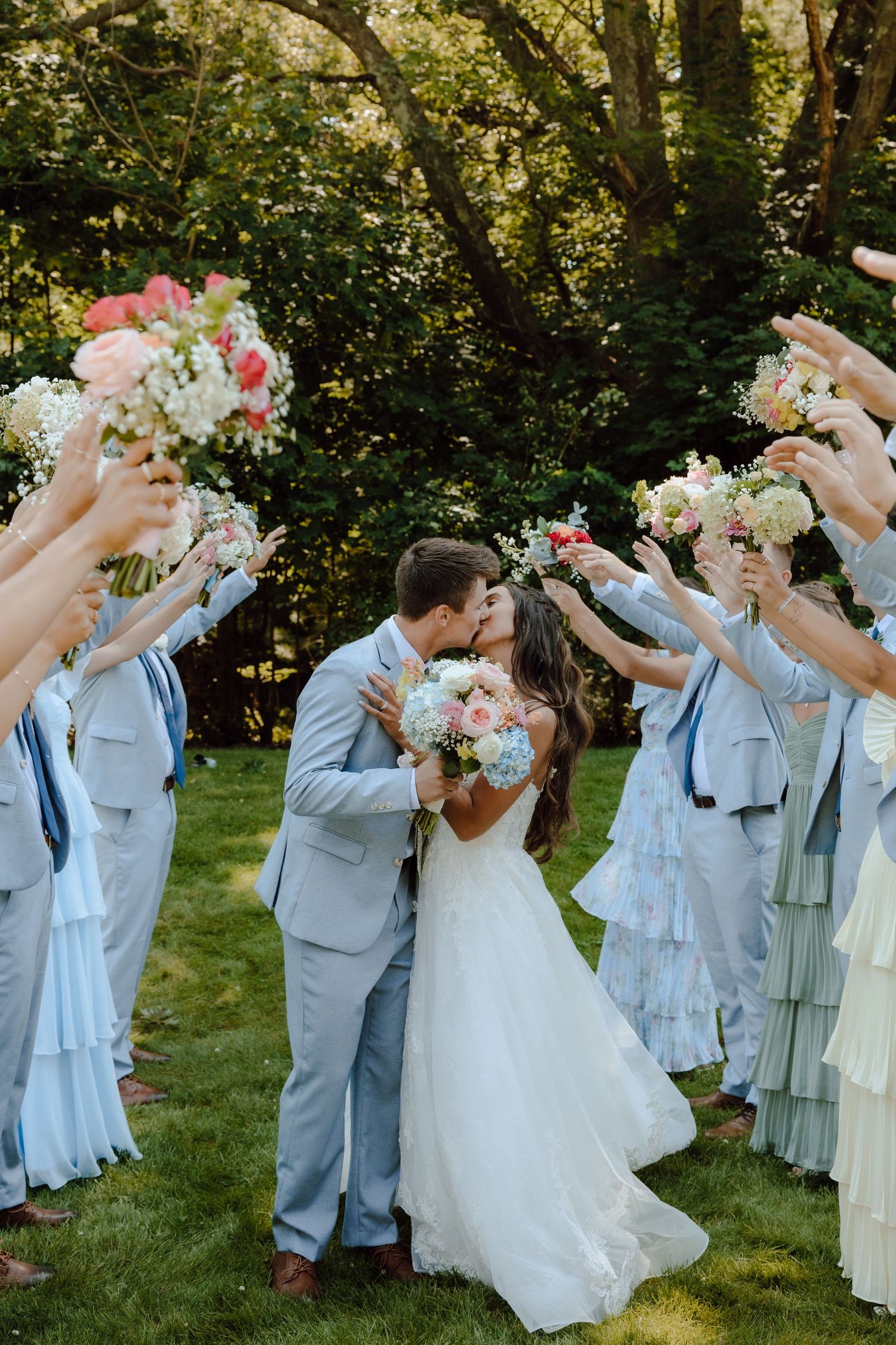 Bride and groom kissing while wedding party raises floral bouquets in a garden ceremony aisle.