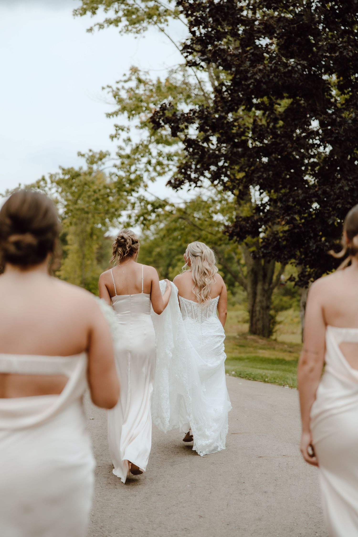 A bride in a white gown walks with bridesmaids in ivory dresses along a tree-lined path at an outdoor wedding.