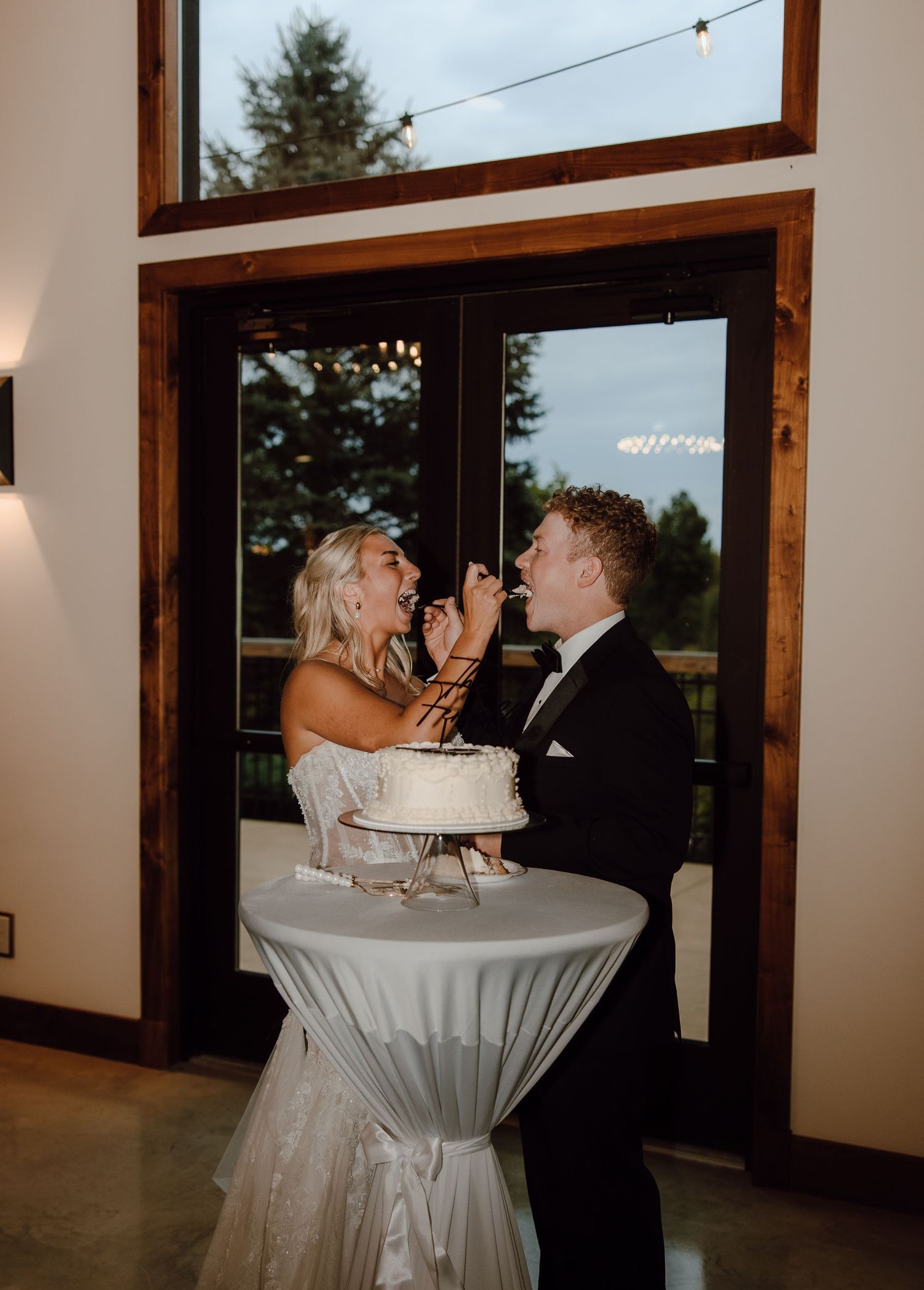 Bride and groom laughing while cutting white wedding cake at elegant indoor reception venue.