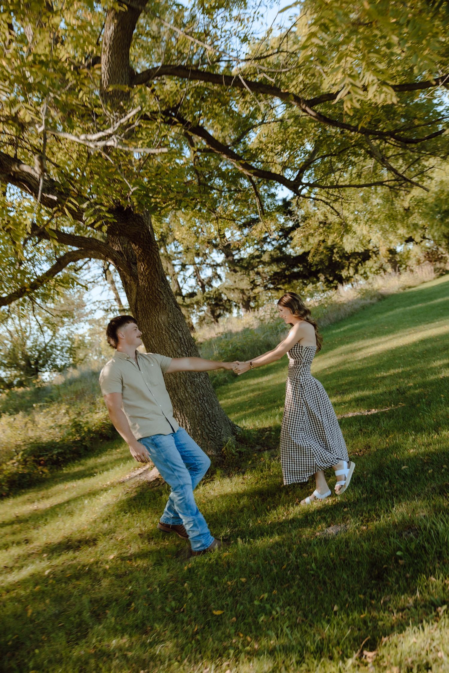 Couple holding hands and dancing under a large tree on a sunny green hillside in a romantic outdoor setting.