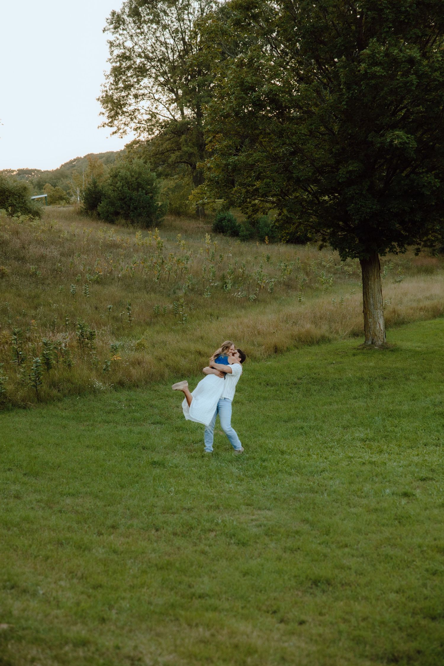 Couple embracing outdoors on lush green grass field with trees in background during golden hour.