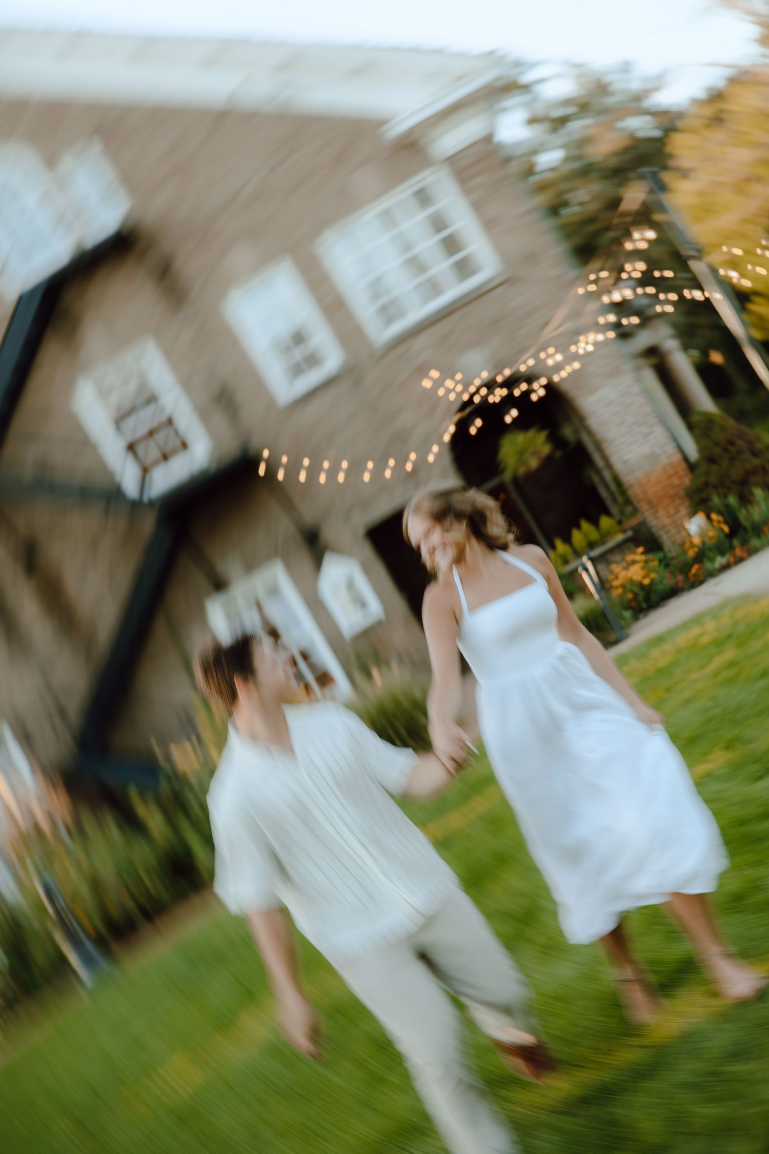 Two brides in white dresses hold hands and dance joyfully on grass outside a brick venue with string lights.