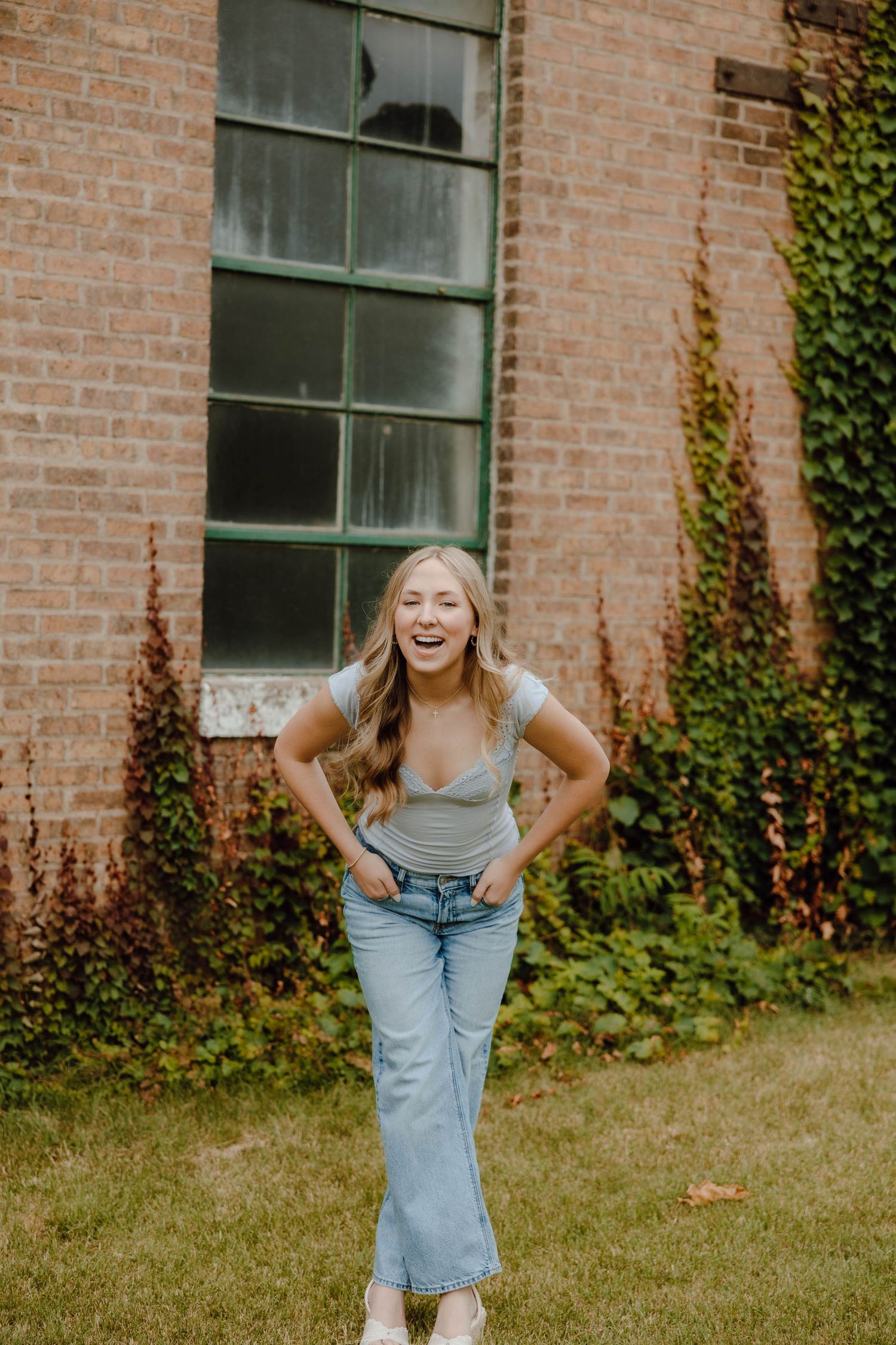 Young woman laughing in light blue jeans and gray top, posing outdoors near brick building with ivy.