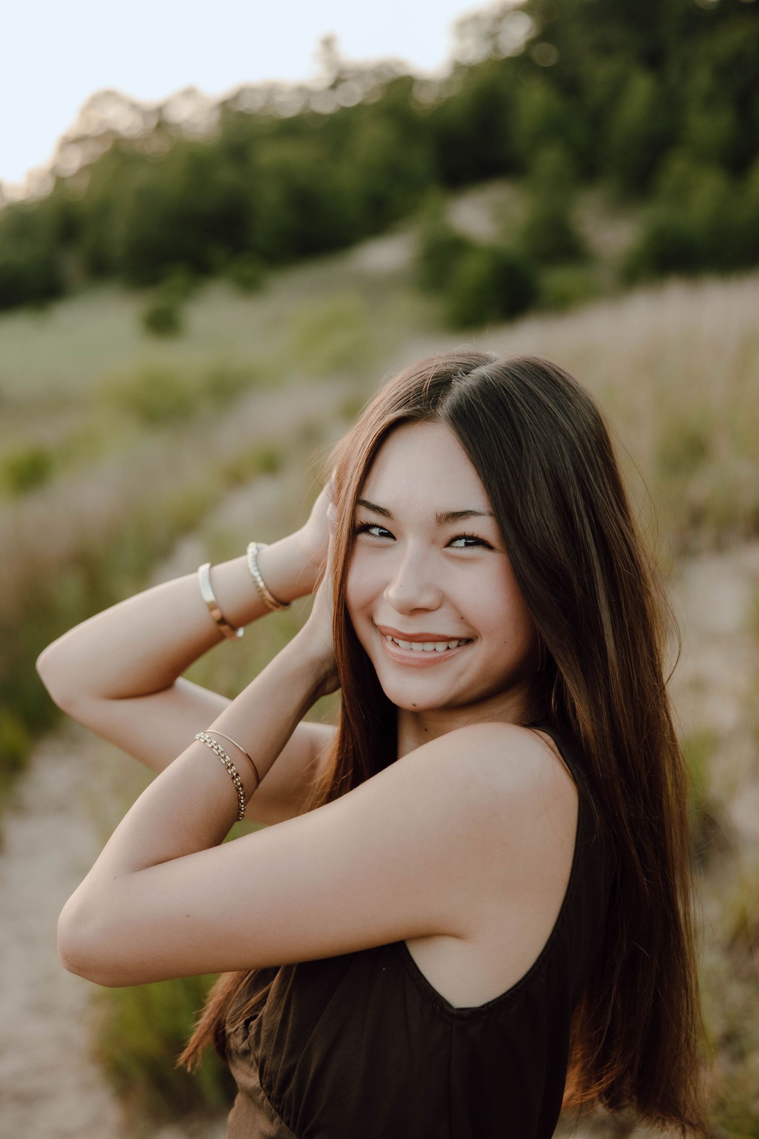 Smiling teenage girl with braces and long brown hair poses outdoors in a black top, wearing gold bracelets.