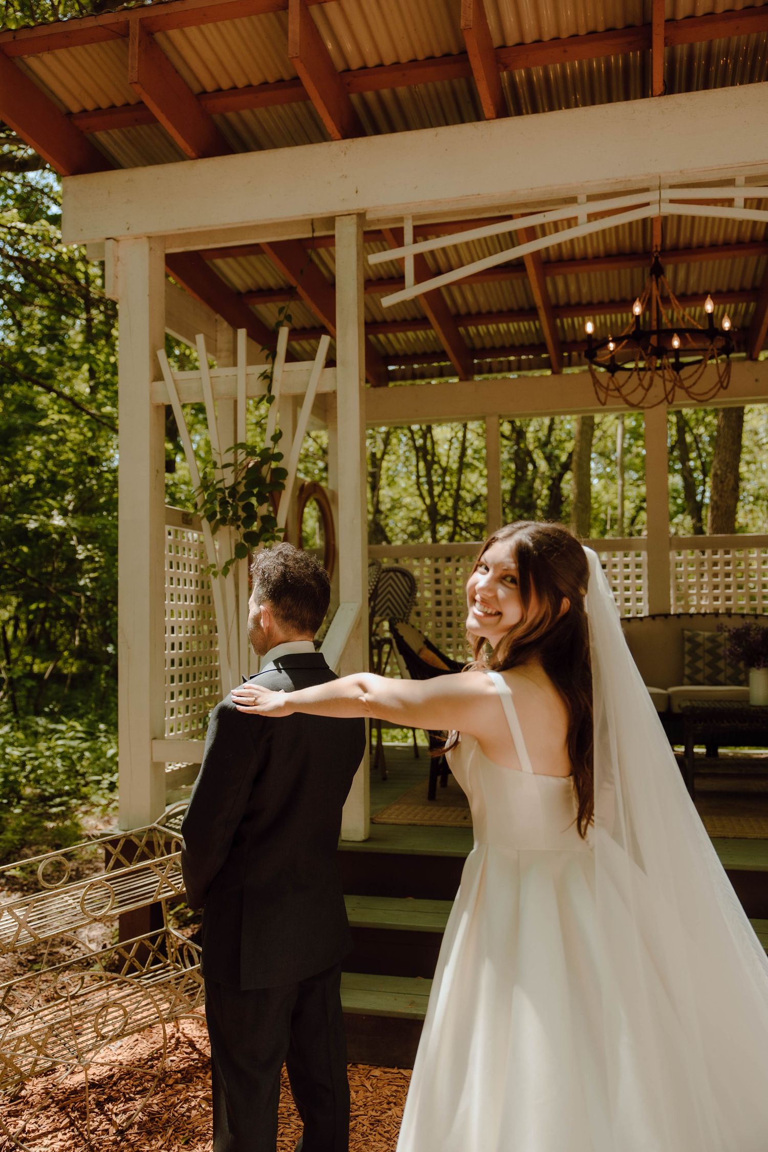 Bride in white gown and veil smiles back at camera during first look moment with groom at outdoor wedding venue.