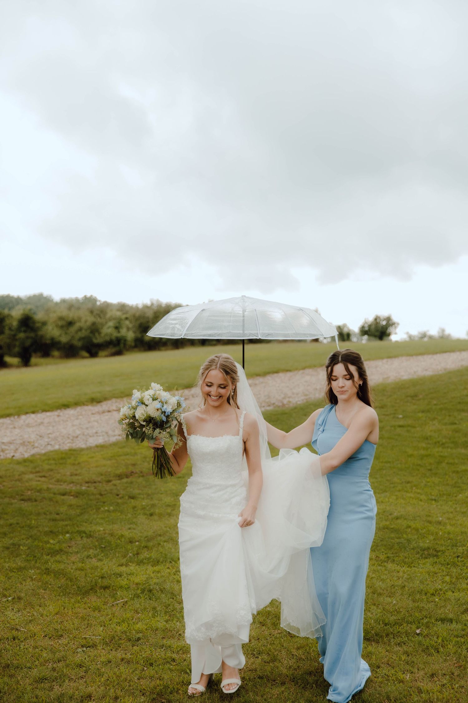 Bridesmaid in blue gown holds bride's train under umbrella on cloudy wedding day outdoors.