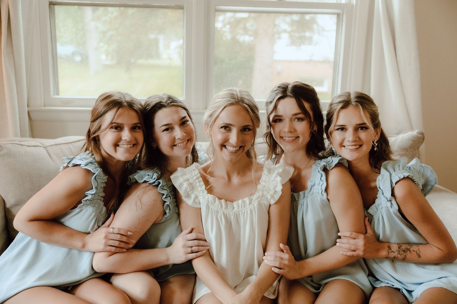 Bride in white surrounded by four bridesmaids in light blue dresses, smiling together on a couch.