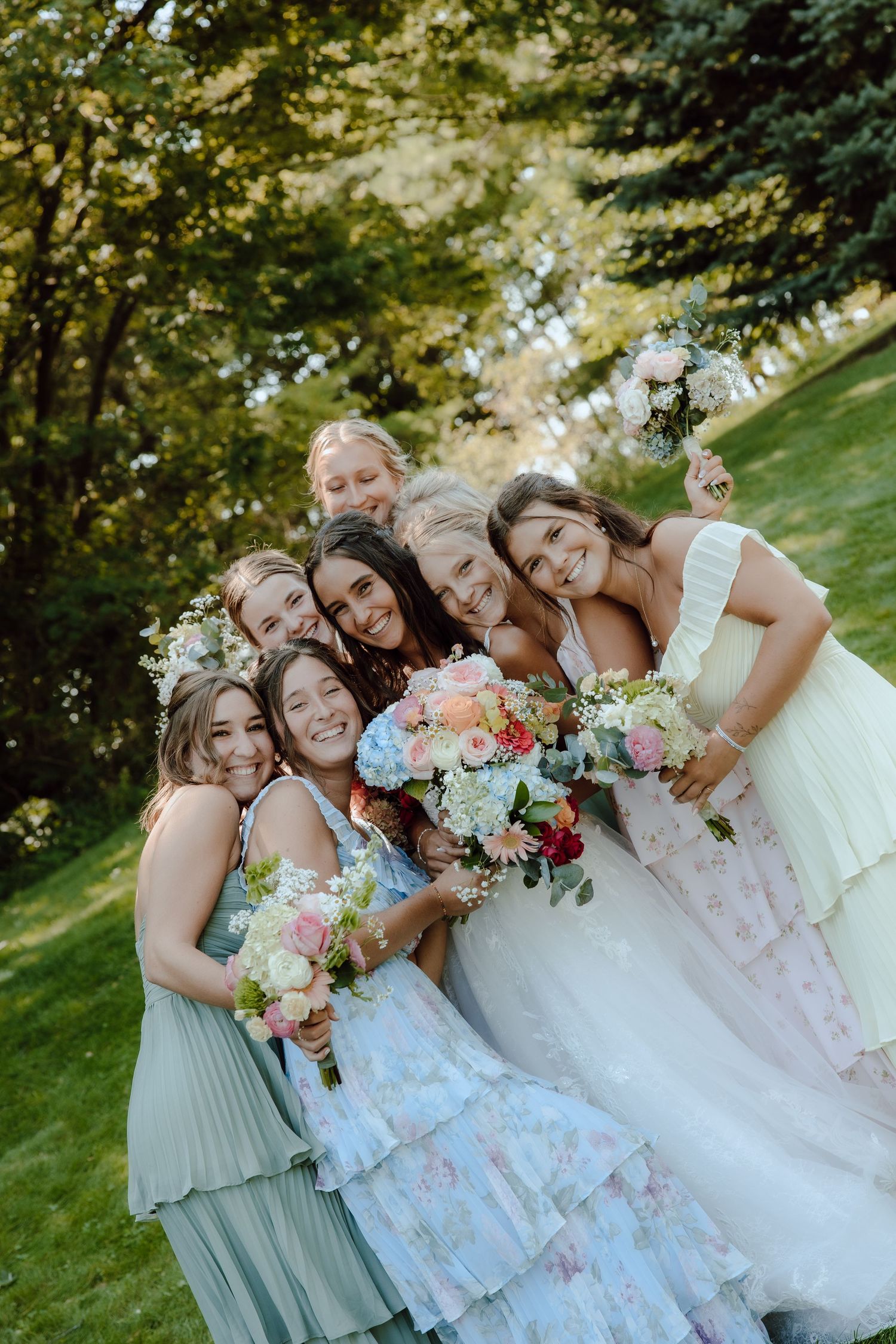 Bride and bridesmaids in colorful dresses hugging and laughing outdoors with bouquets.