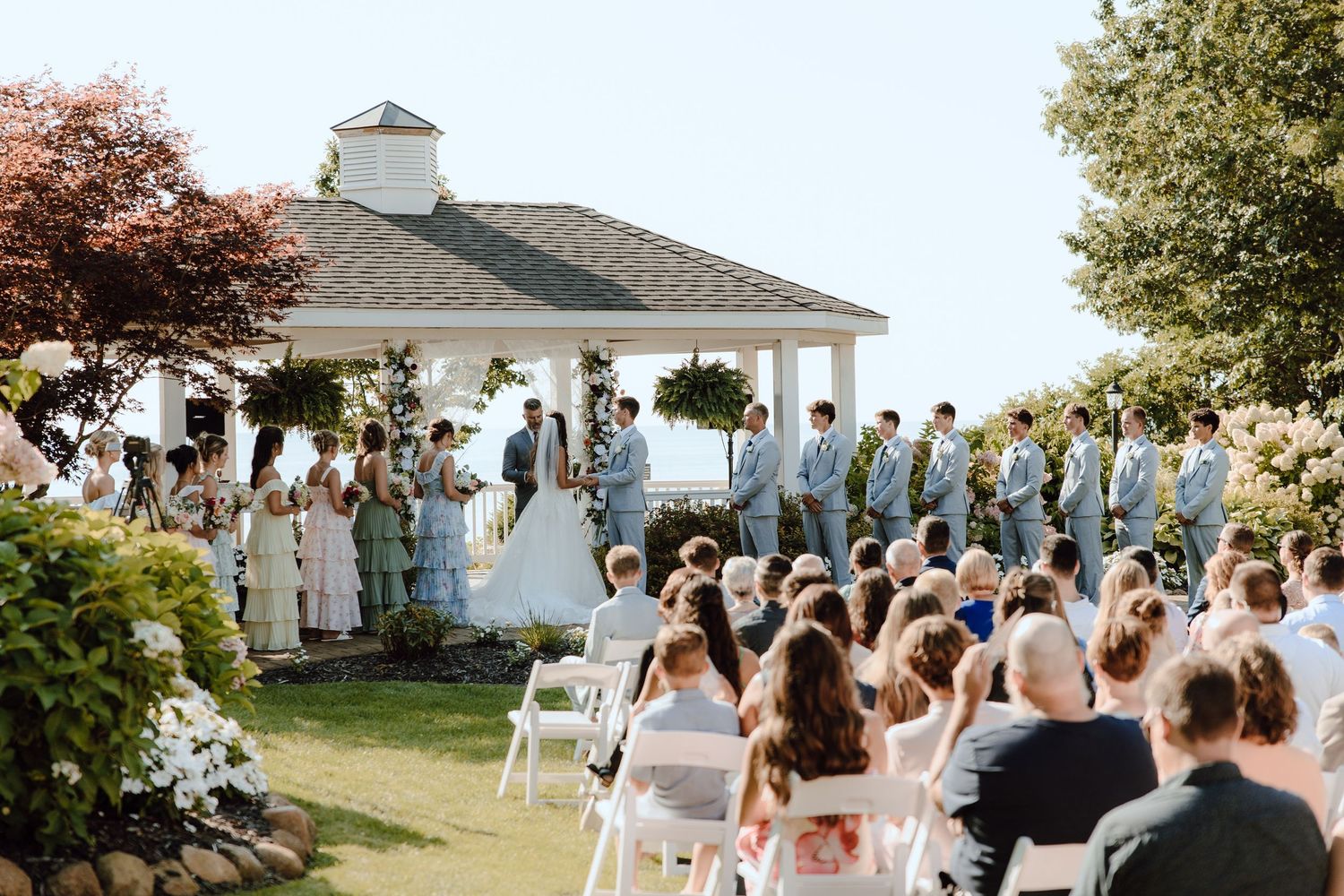 Outdoor wedding ceremony on a lawn with white chairs, bridal party, and a white gazebo backdrop on a sunny day.