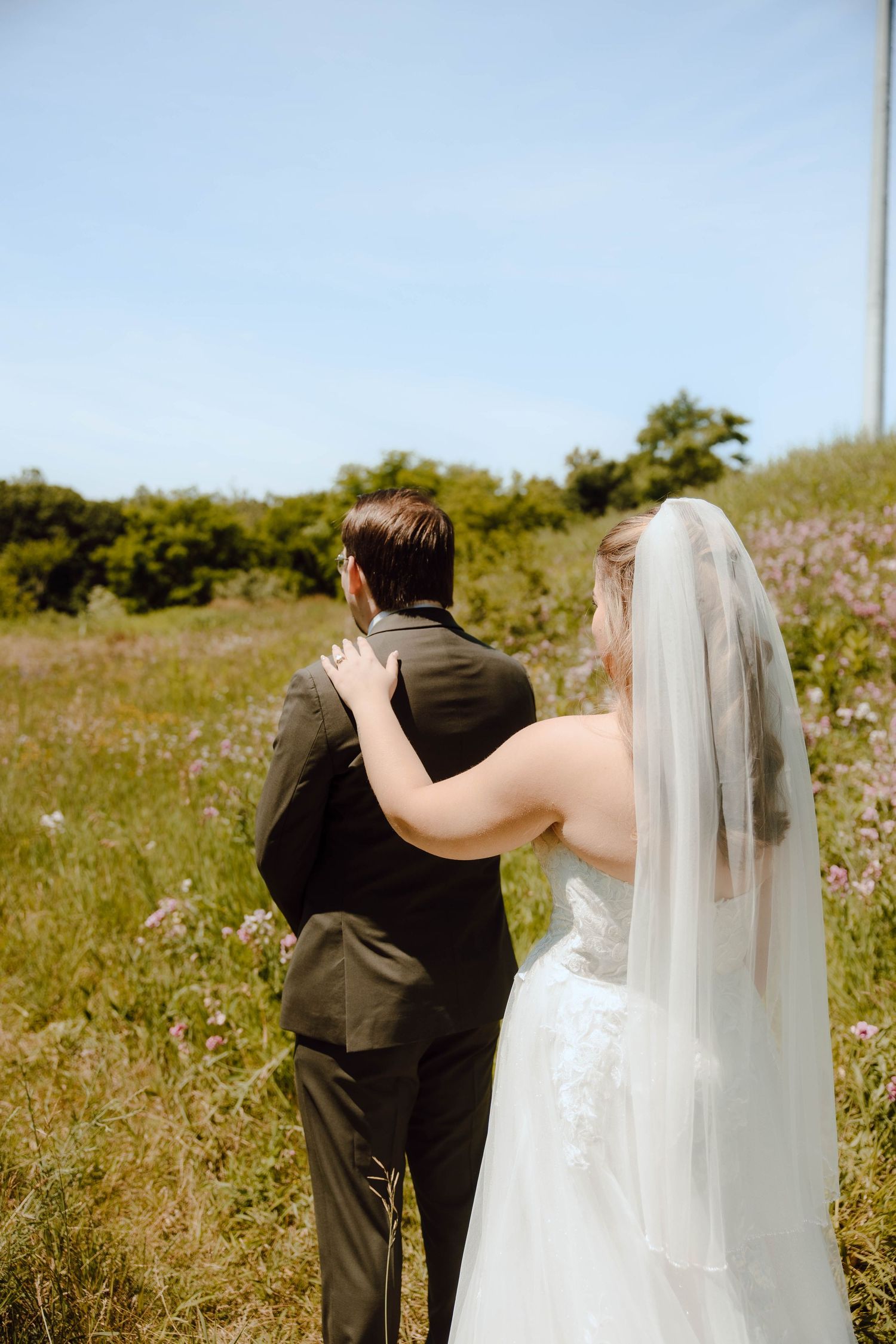 A bride in a white gown and veil places her hand on a groom in a dark suit as they look out over a wildflower field.