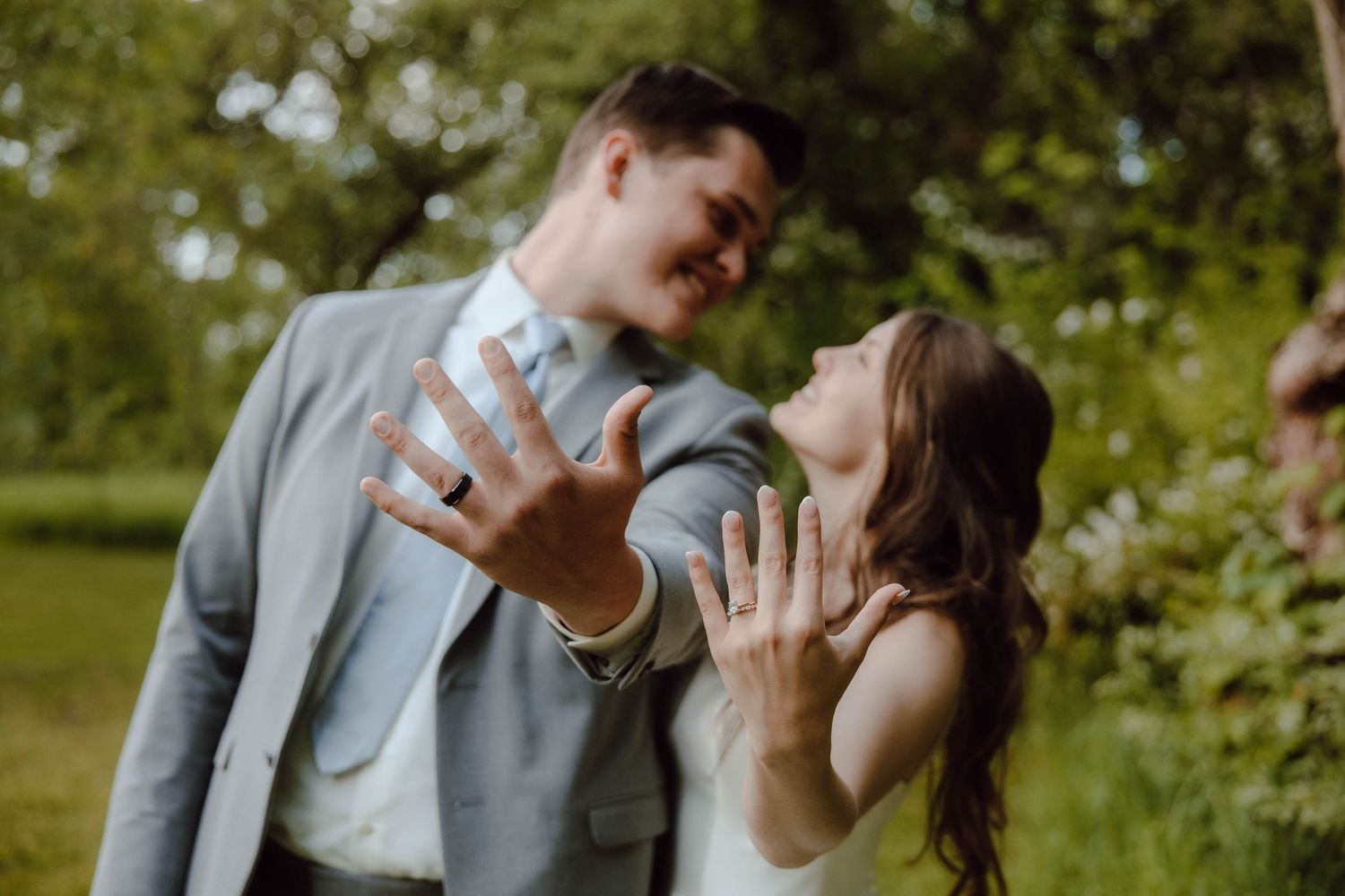 Newlyweds joyfully show off wedding rings outdoors, smiling couple in gray suit and white dress.