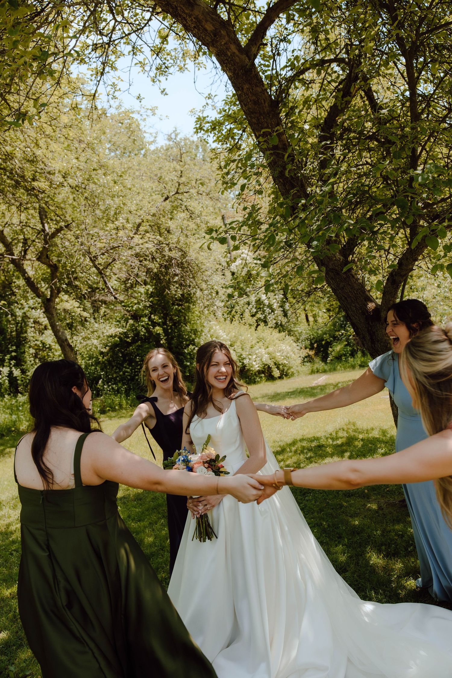 Bride in white gown holding bouquet laughing with bridesmaids in colorful dresses outdoors under green trees.