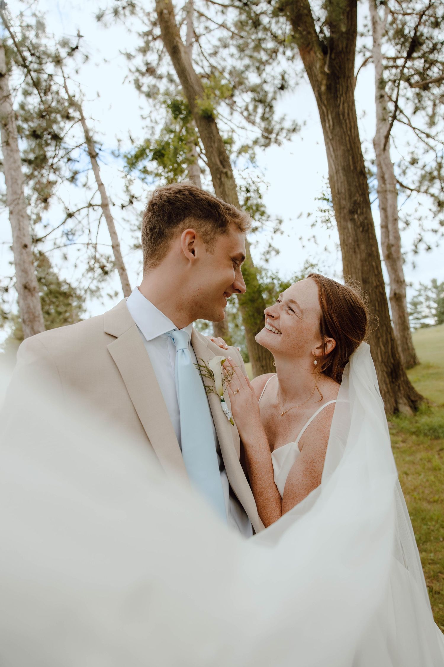 Bride and groom share a joyful moment outdoors, surrounded by tall trees, as she laughs in a white gown and veil.