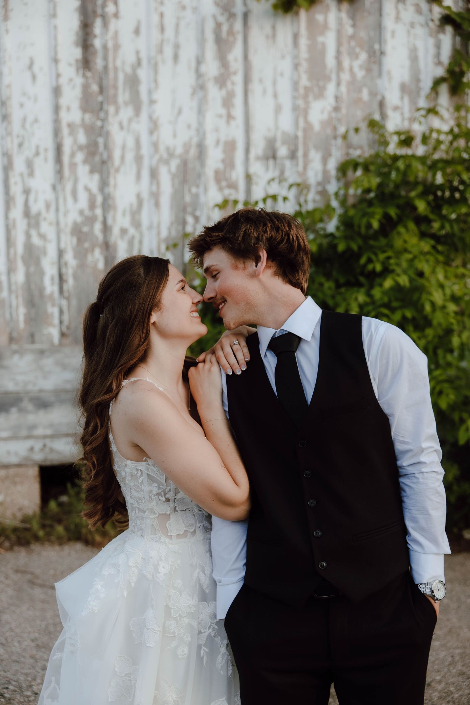 A bride and groom share a romantic almost-kiss moment outdoors, dressed in wedding attire against a rustic white barn wall.