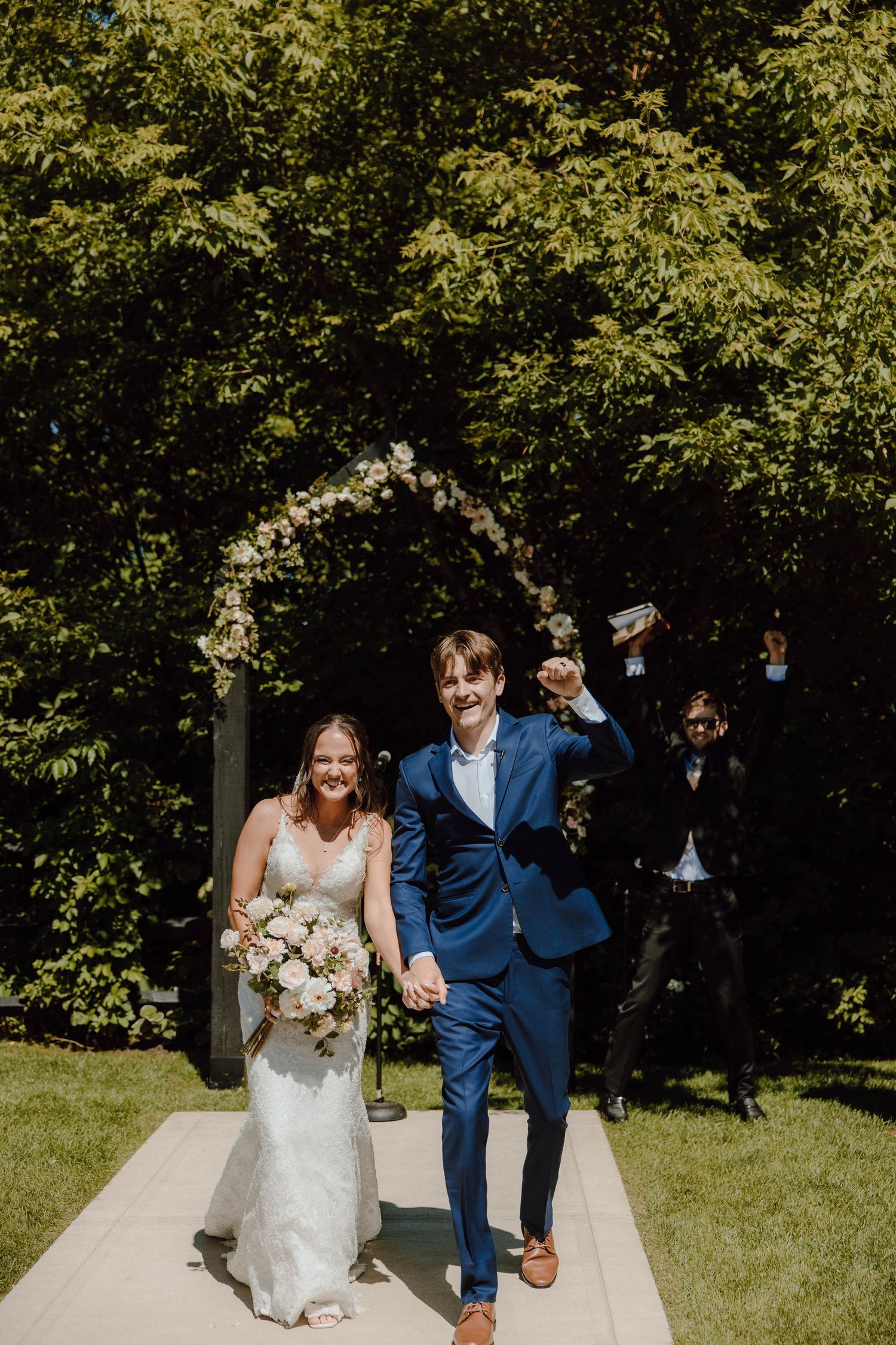 Newlyweds walk hand in hand down aisle after ceremony, groom in blue suit raises fist in celebration outdoors.