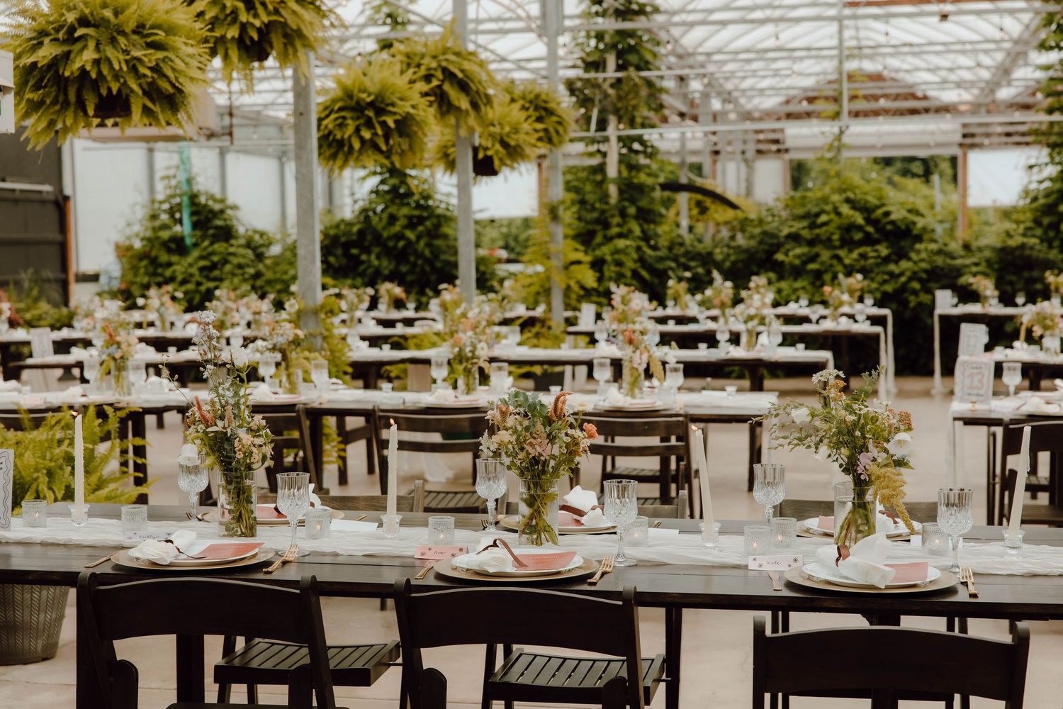 Elegant greenhouse wedding reception with long tables, floral centerpieces, dark chairs, and lush hanging plants overhead.