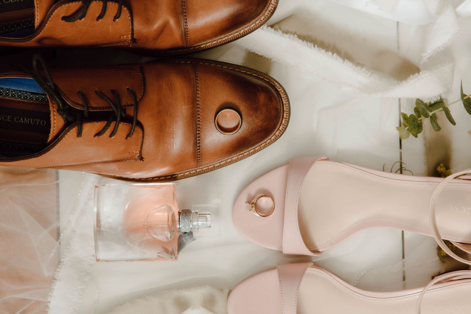 Brown leather dress shoes and pink heels with wedding rings and perfume on white fur, bridal flat lay.
