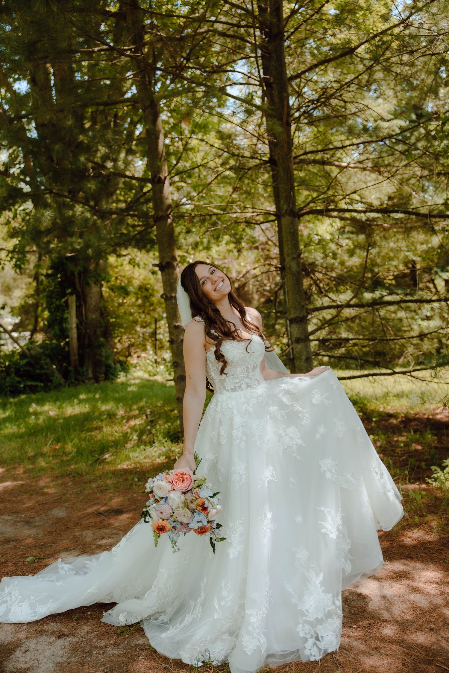 Smiling bride in a white lace ball gown holding a floral bouquet, posing outdoors in a sunlit forest setting.
