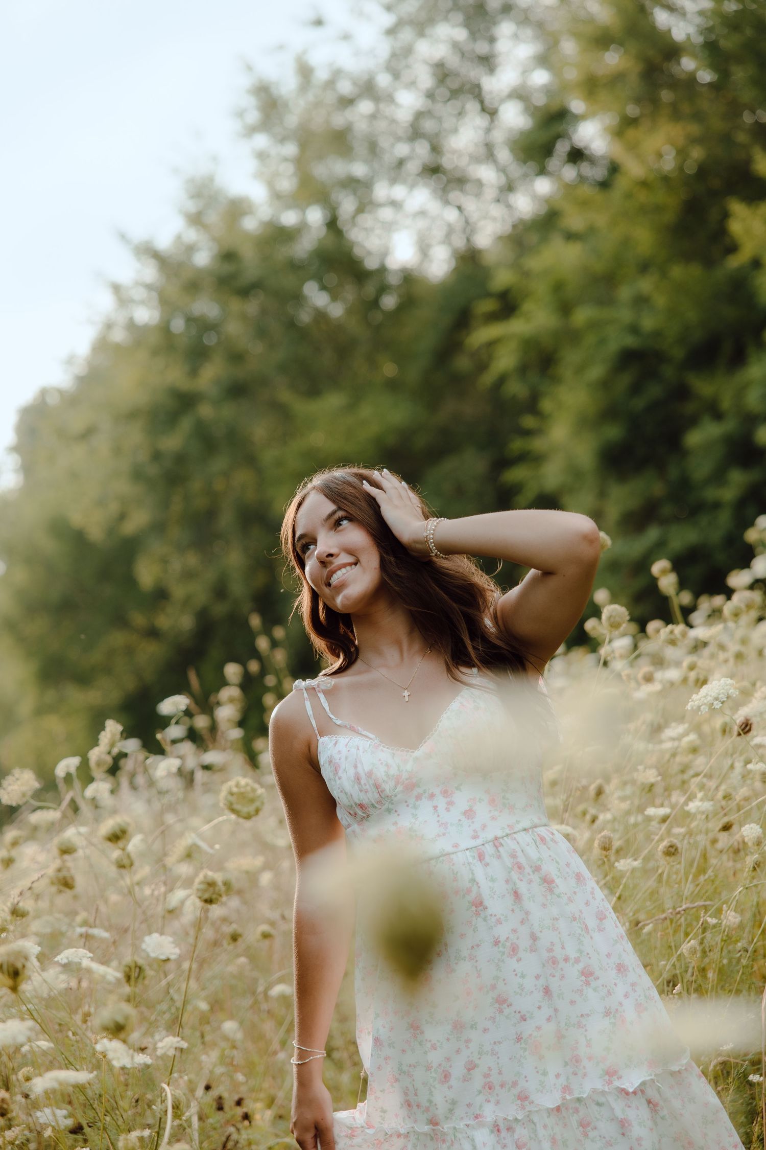 Smiling young woman in white floral sundress standing among wildflowers in a sun-dappled meadow.