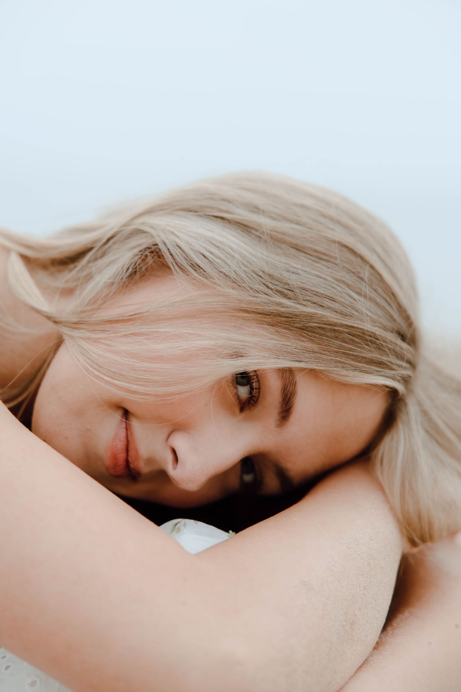 Smiling blonde woman resting her head on her arms, looking up with a serene expression against a light background.