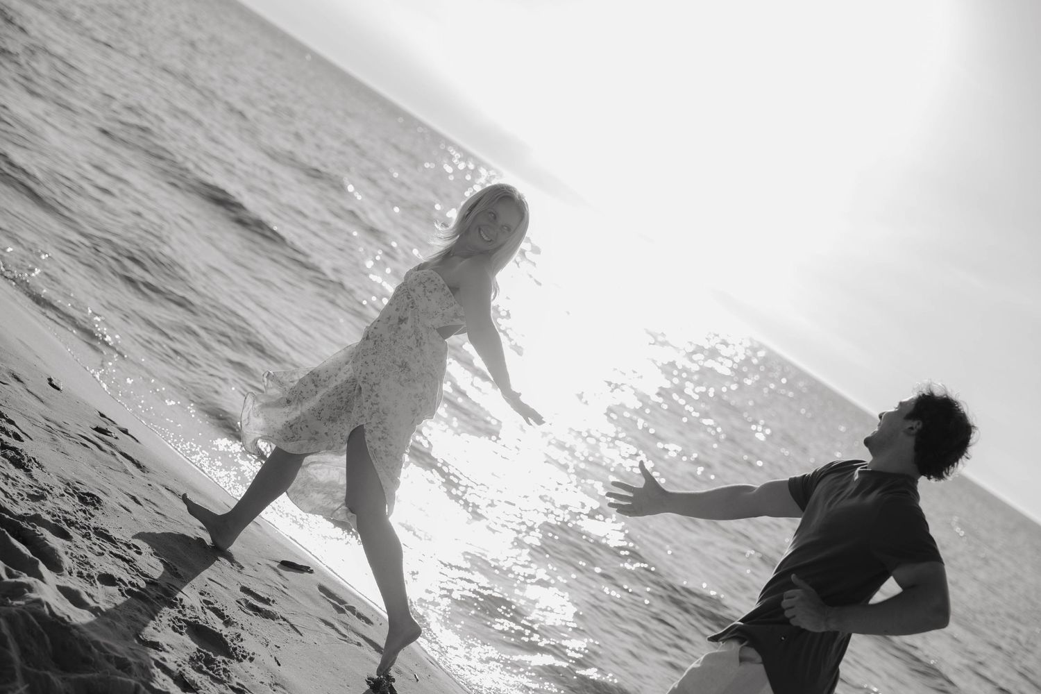 Black and white photo of two people playing near ocean waves on a sunny beach.
