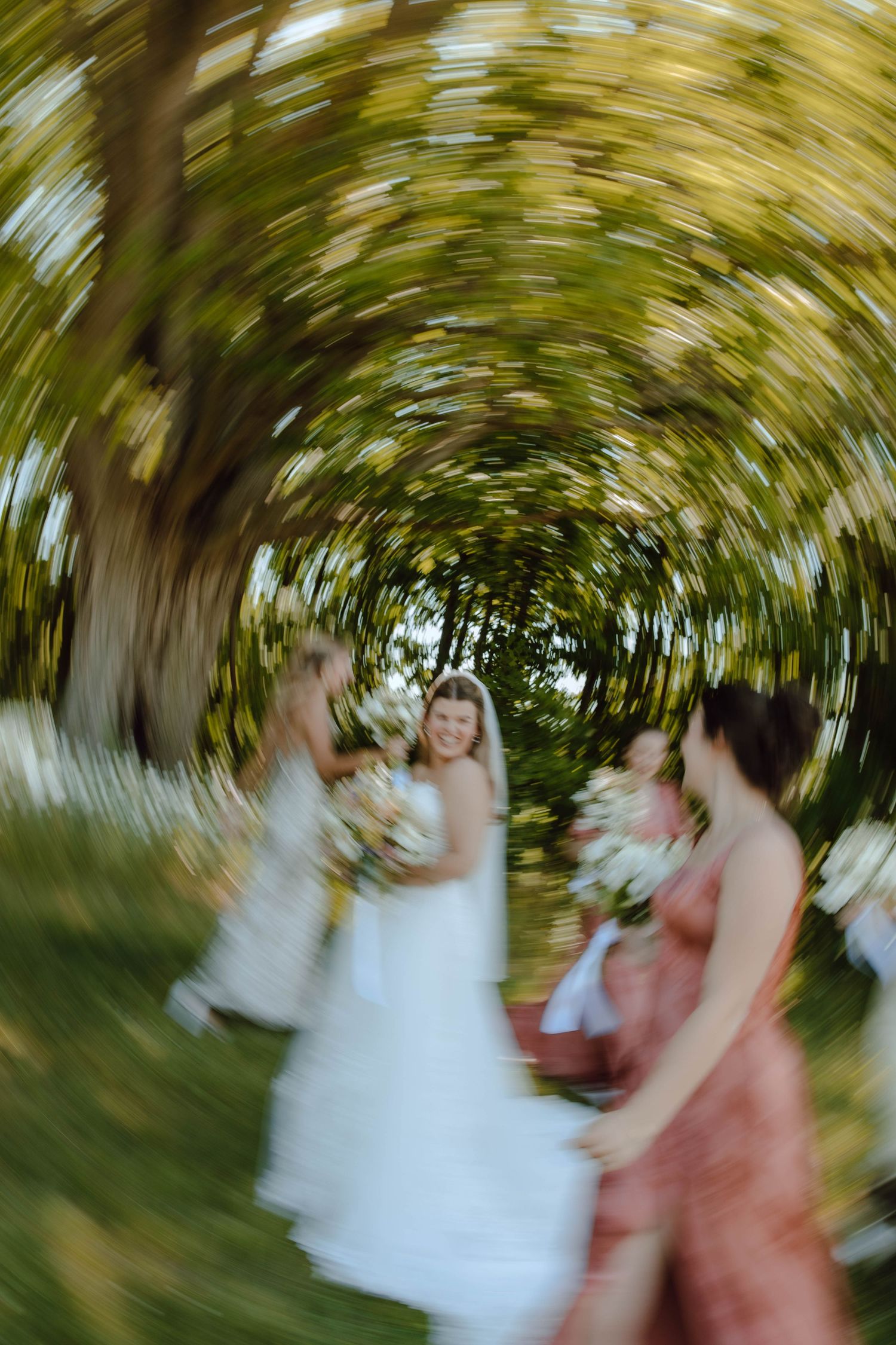 Creative spin blur wedding photo of bride and bridesmaids celebrating under lush green trees.