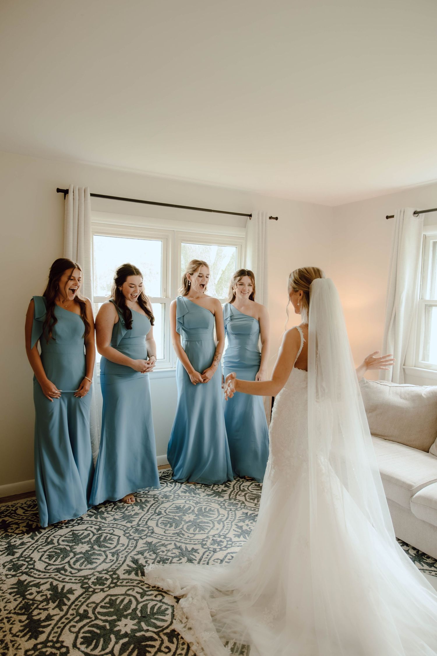 Bride reveals wedding dress to four bridesmaids in dusty blue gowns in a bright white getting-ready room.