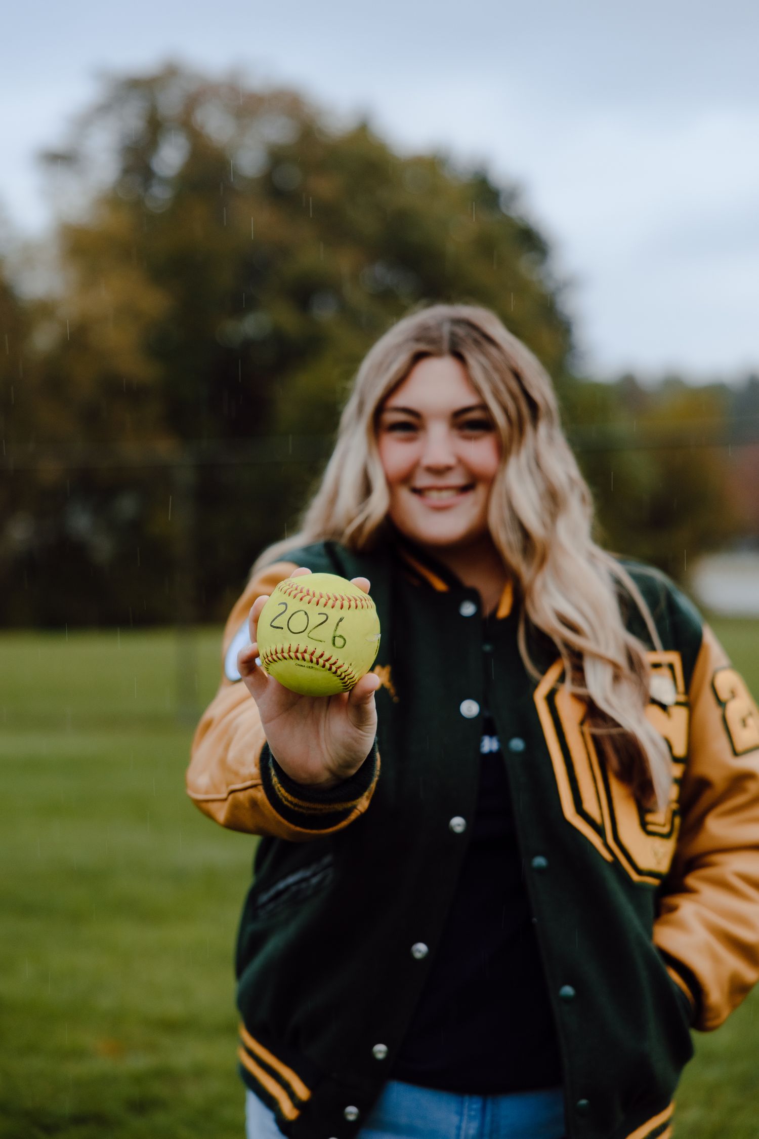 A varsity letterman jacket and softball being held up against an autumn field background.