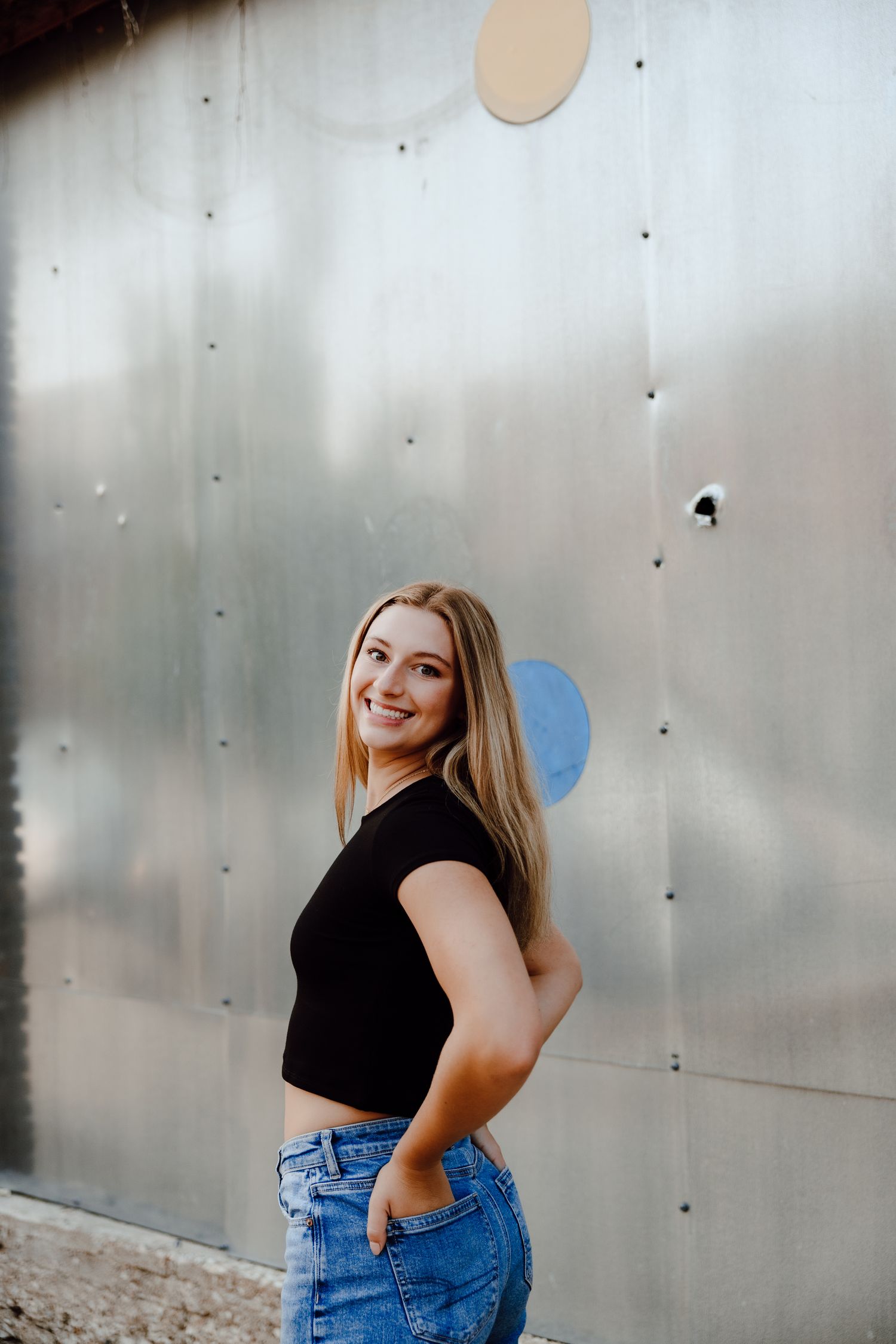 Person in black top and jeans poses playfully against metallic wall.
