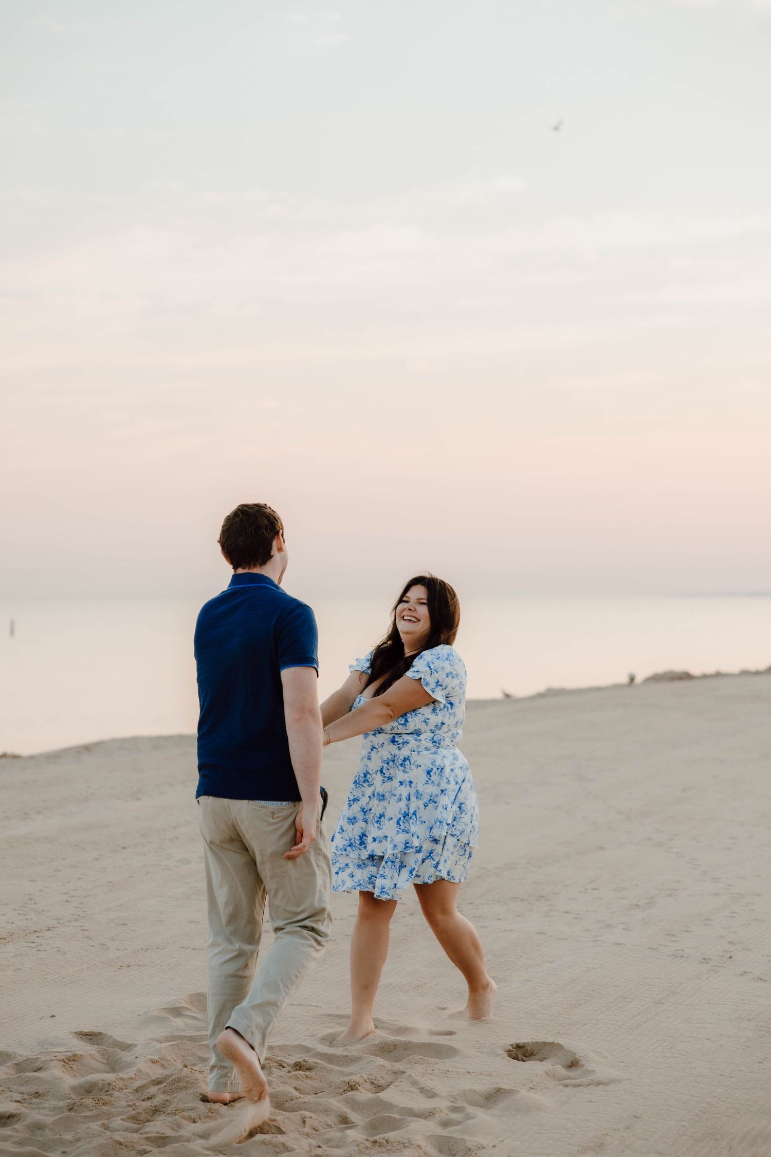 A couple dances barefoot on a sandy beach at sunset, the woman in a floral dress laughing joyfully.