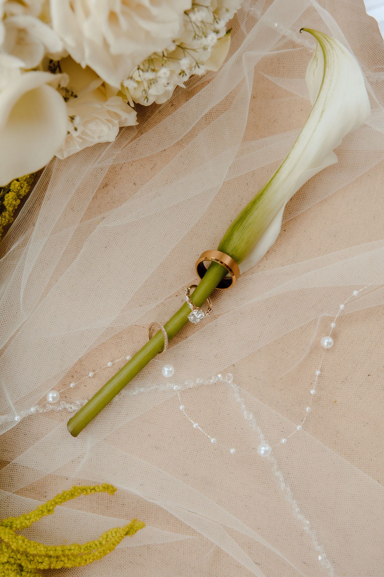 Wedding rings resting on a white calla lily stem atop a bridal veil with pearl details.