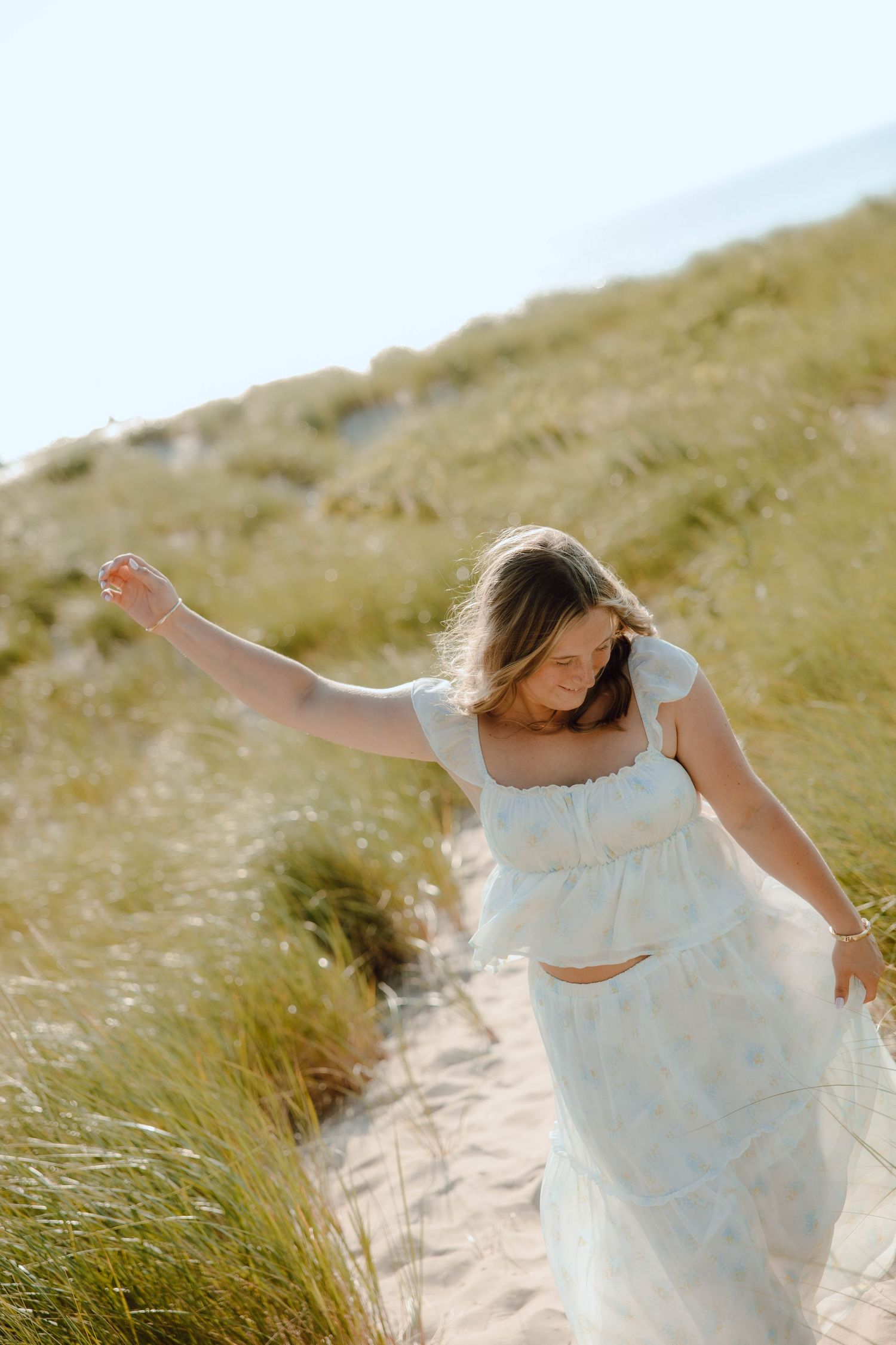 Young woman in white dress walking through sandy beach dunes with tall grass on a sunny day.