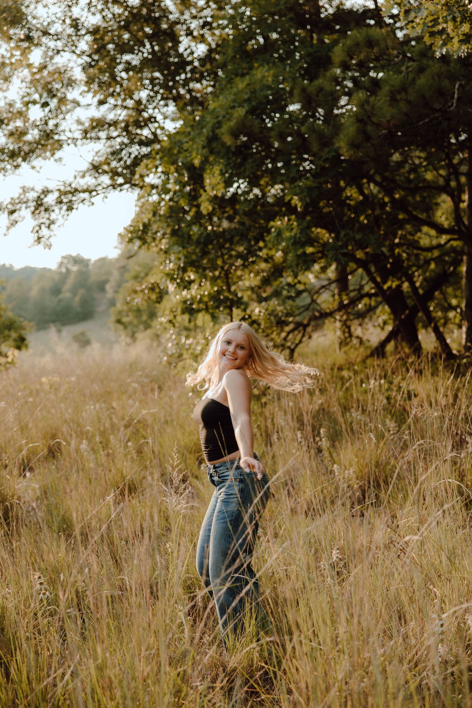 Blonde woman in black top and jeans standing in golden tall grass field with trees in background.